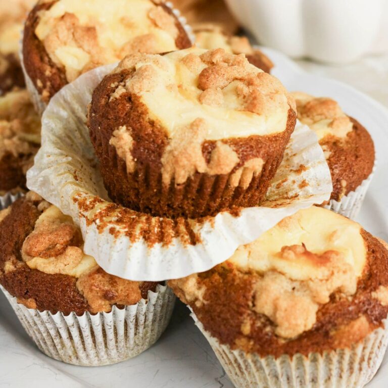 A close-up of several muffins with crumbly topping and cream cheese filling, some stacked on a white plate, with one muffin partially unwrapped.