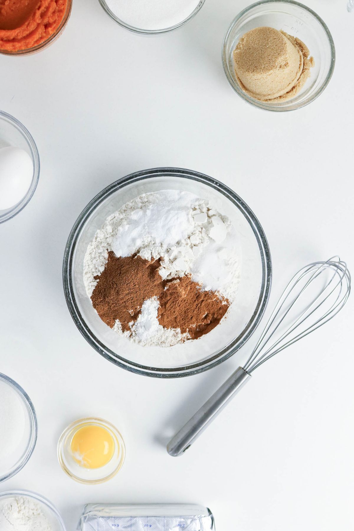 A bowl with flour, baking powder, and cinnamon sits on a white surface surrounded by a whisk, eggs, sugars, and other baking ingredients in separate bowls.