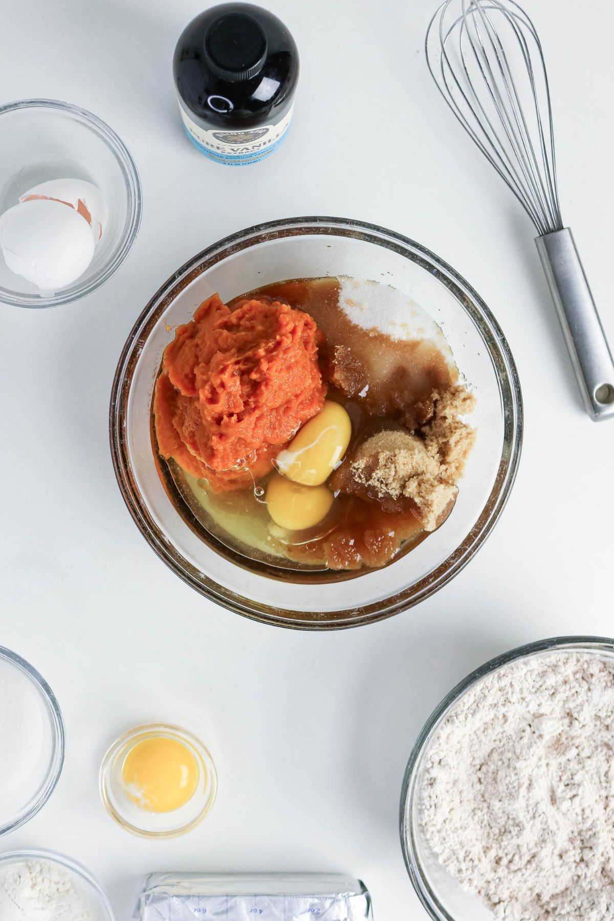 A glass bowl with pumpkin puree, eggs, sugar, and vanilla surrounded by flour, sugar, an egg yolk, a whisk, vanilla extract, and an egg on a white surface.