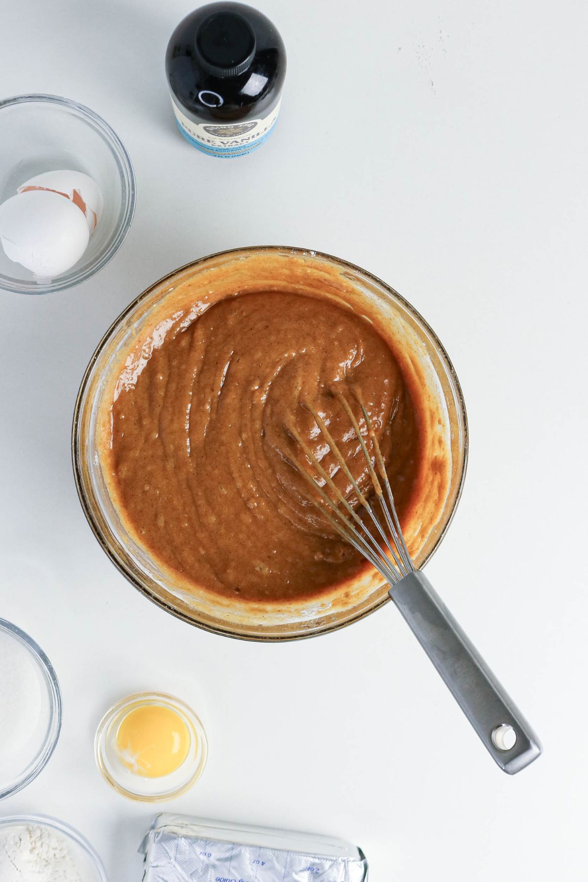 A glass bowl with a whisk mixing brown batter, surrounded by eggs, vanilla extract, sugar, flour, and butter on a white surface.