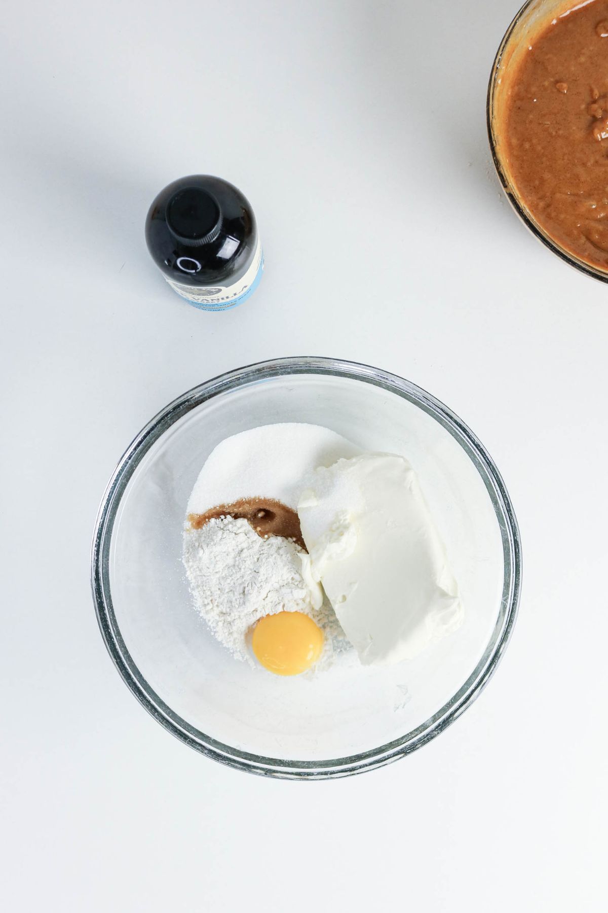 A glass bowl with flour, sugar, cream cheese, an egg yolk, and vanilla extract on a white surface; nearby are a bottle and a bowl with a brown mixture.