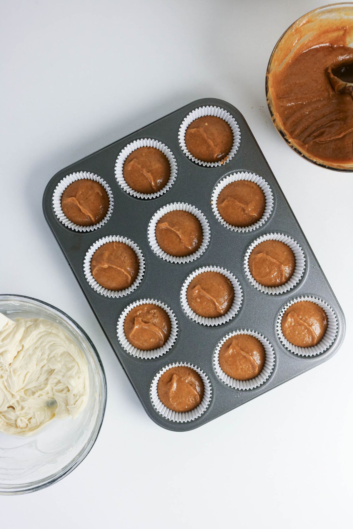 A muffin tray with twelve paper liners, each filled with brown batter, sits on a white surface next to bowls of white and brown batter.