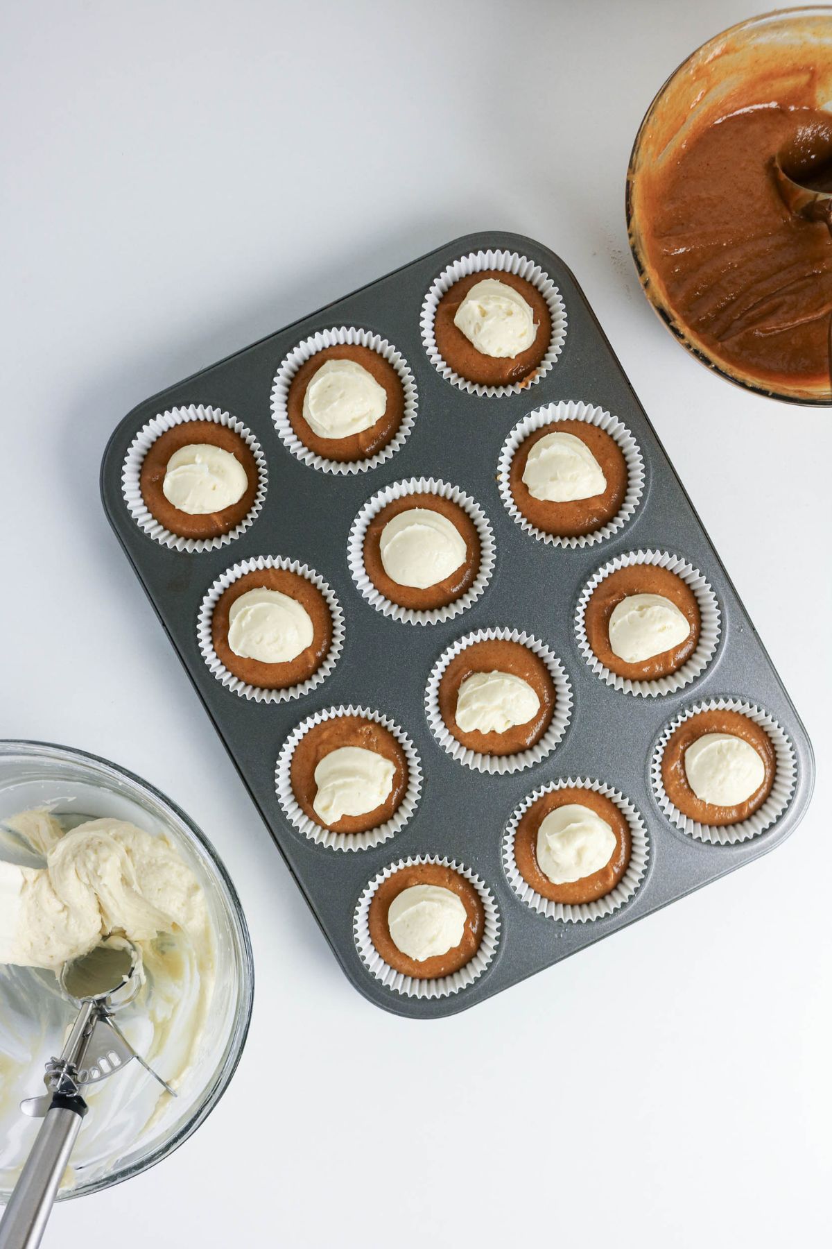 Muffin tin with 12 cupcake liners filled with brown batter and a dollop of white cream in each, with bowls of batters and scoop nearby on a white surface.