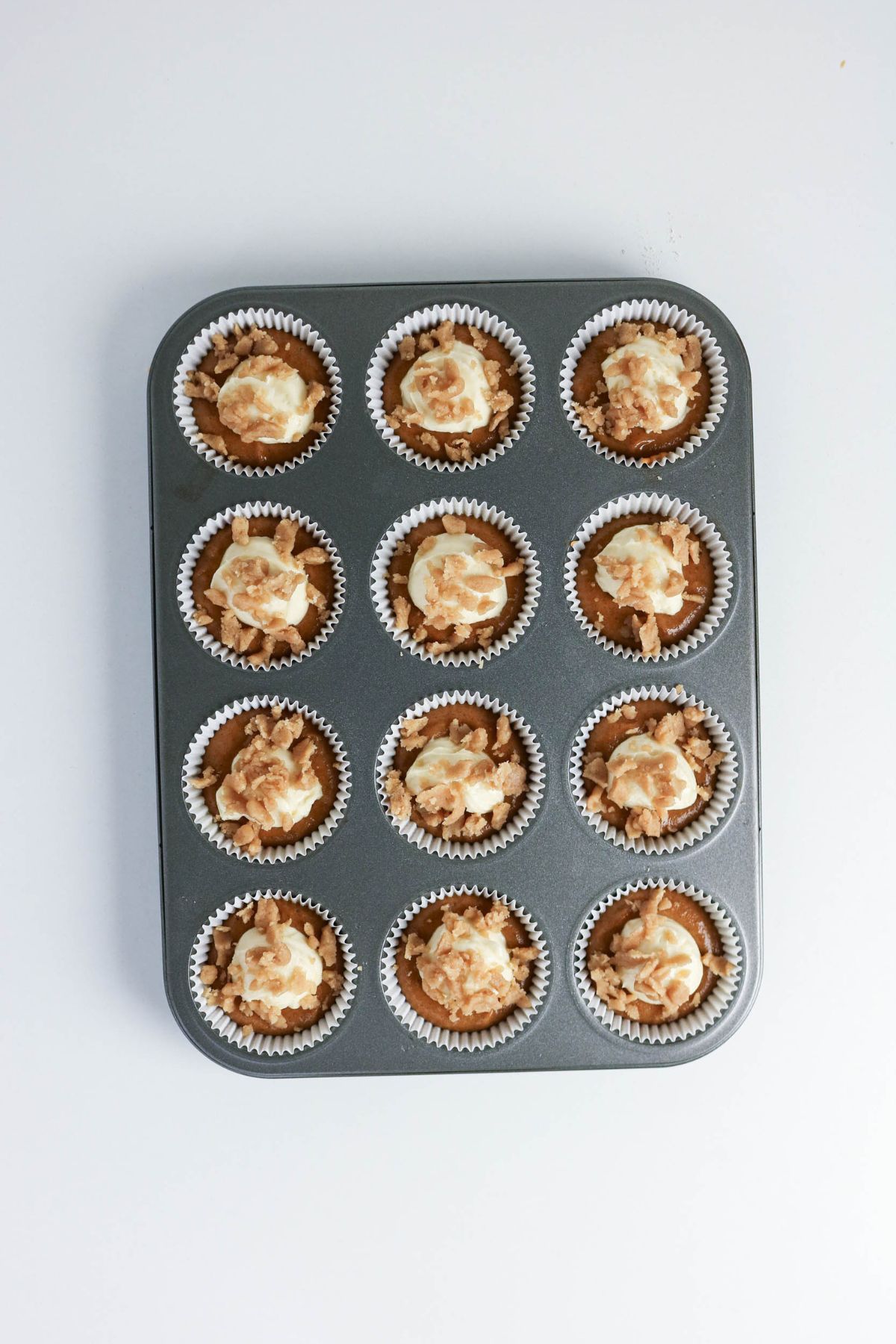 A muffin tray holds twelve baked cupcakes with white paper liners and swirled topping, viewed from above on a plain white background.