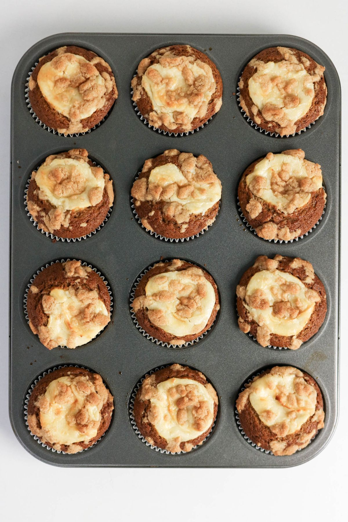 A muffin tin with twelve baked muffins, each topped with a crumbly and creamy swirl, viewed from above on a plain white background.