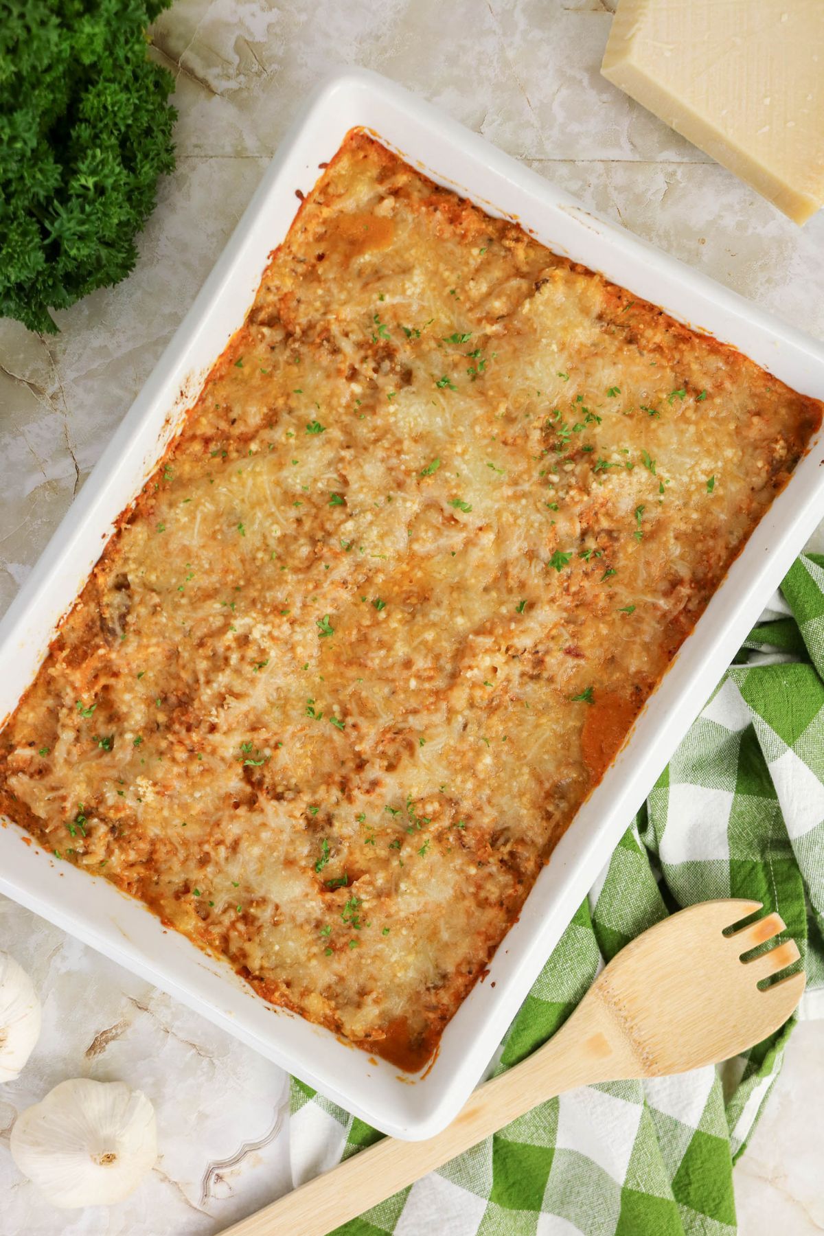 A baked lasagna in a white rectangular dish, topped with melted cheese and herbs, sits on a marble surface beside a green checked towel, wooden spoon, and fresh garlic.