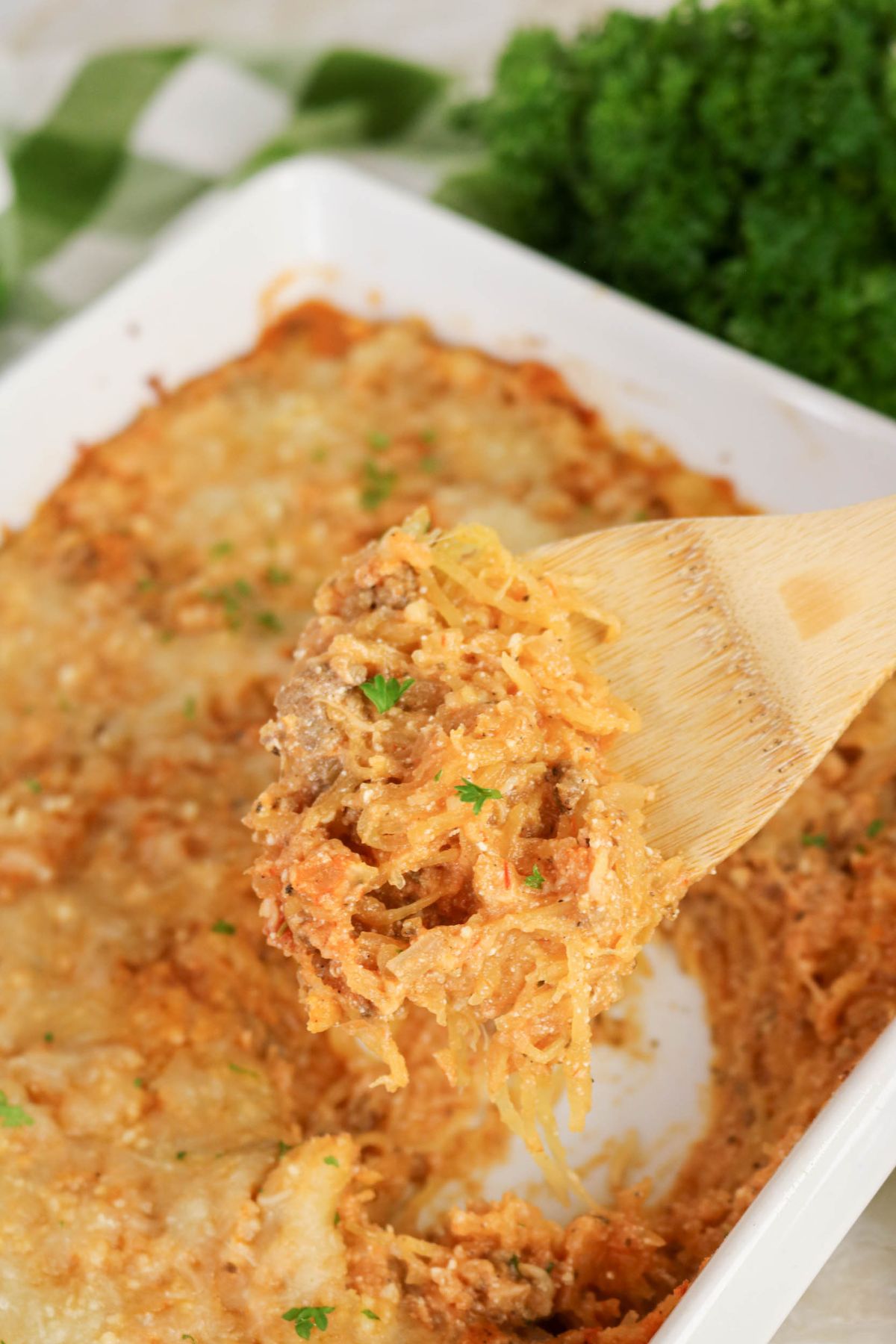 A wooden spoon lifts a serving of baked casserole with melted cheese from a white dish, with parsley garnish visible.