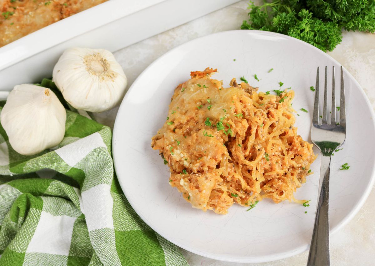 A serving of baked spaghetti topped with melted cheese and parsley on a white plate, with a fork, garlic bulbs, green checkered cloth, and parsley nearby.