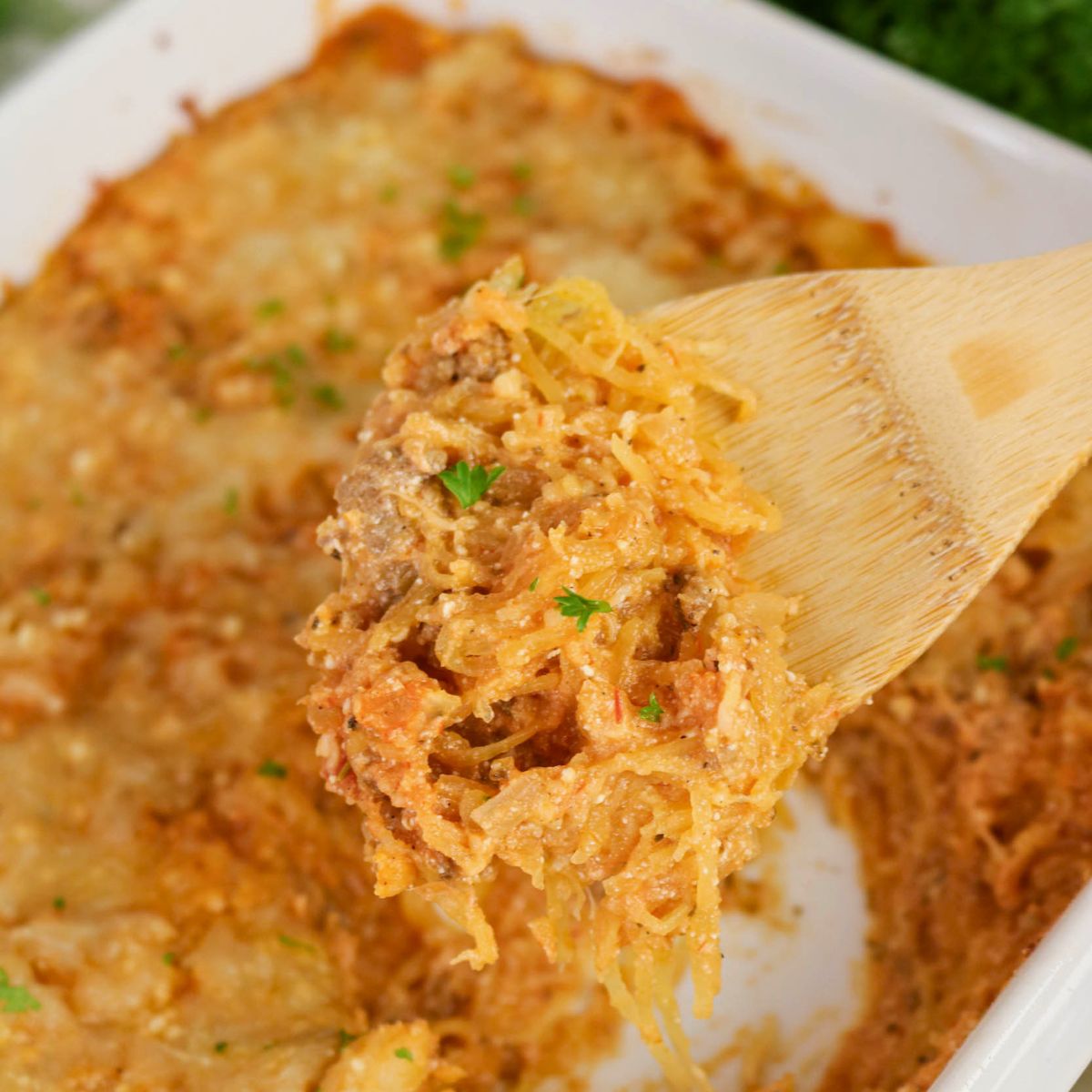 A wooden spoon holds a serving of cheesy baked spaghetti squash casserole above a white baking dish.