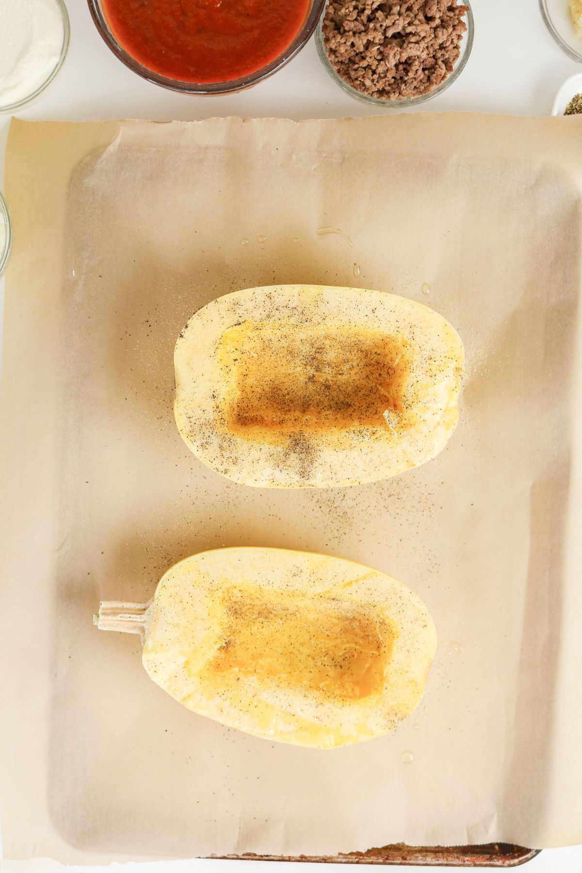 Two spaghetti squash halves face up on parchment paper, seasoned with pepper, surrounded by bowls of tomato sauce, ground meat, and other ingredients.