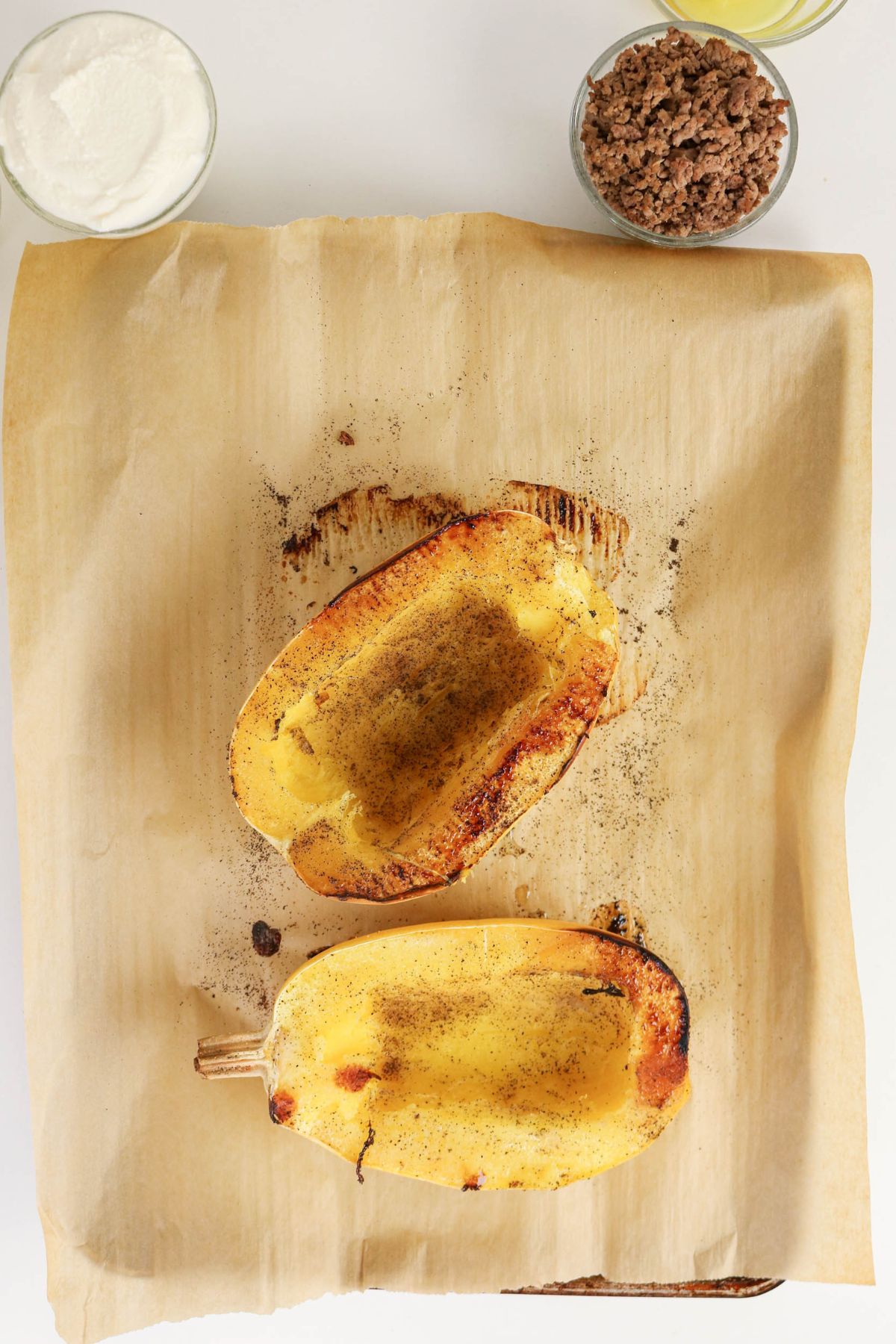 Two roasted spaghetti squash halves on parchment paper, with small bowls of ground meat and white sauce placed above them.