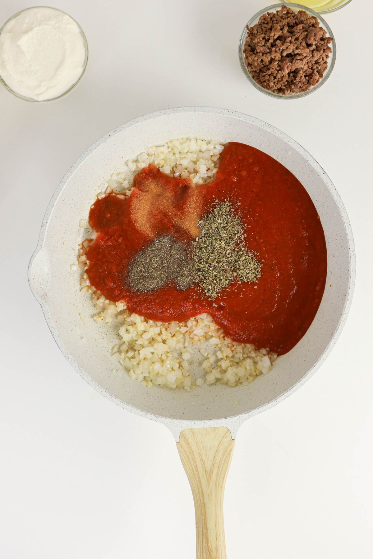 White skillet with chopped onions, tomato sauce, and assorted spices. Nearby bowls contain ricotta cheese and cooked ground meat.