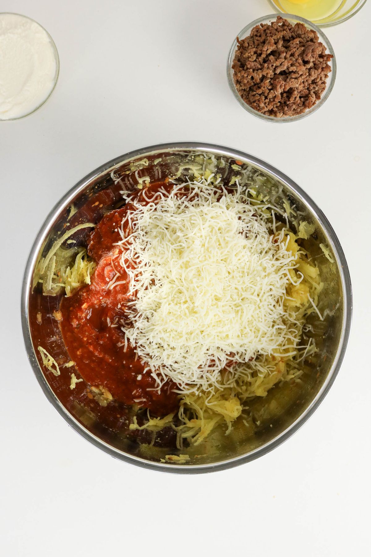 A metal bowl contains shredded cheese, tomato sauce, and spaghetti squash, with bowls of cooked ground meat and ricotta cheese nearby on a white surface.