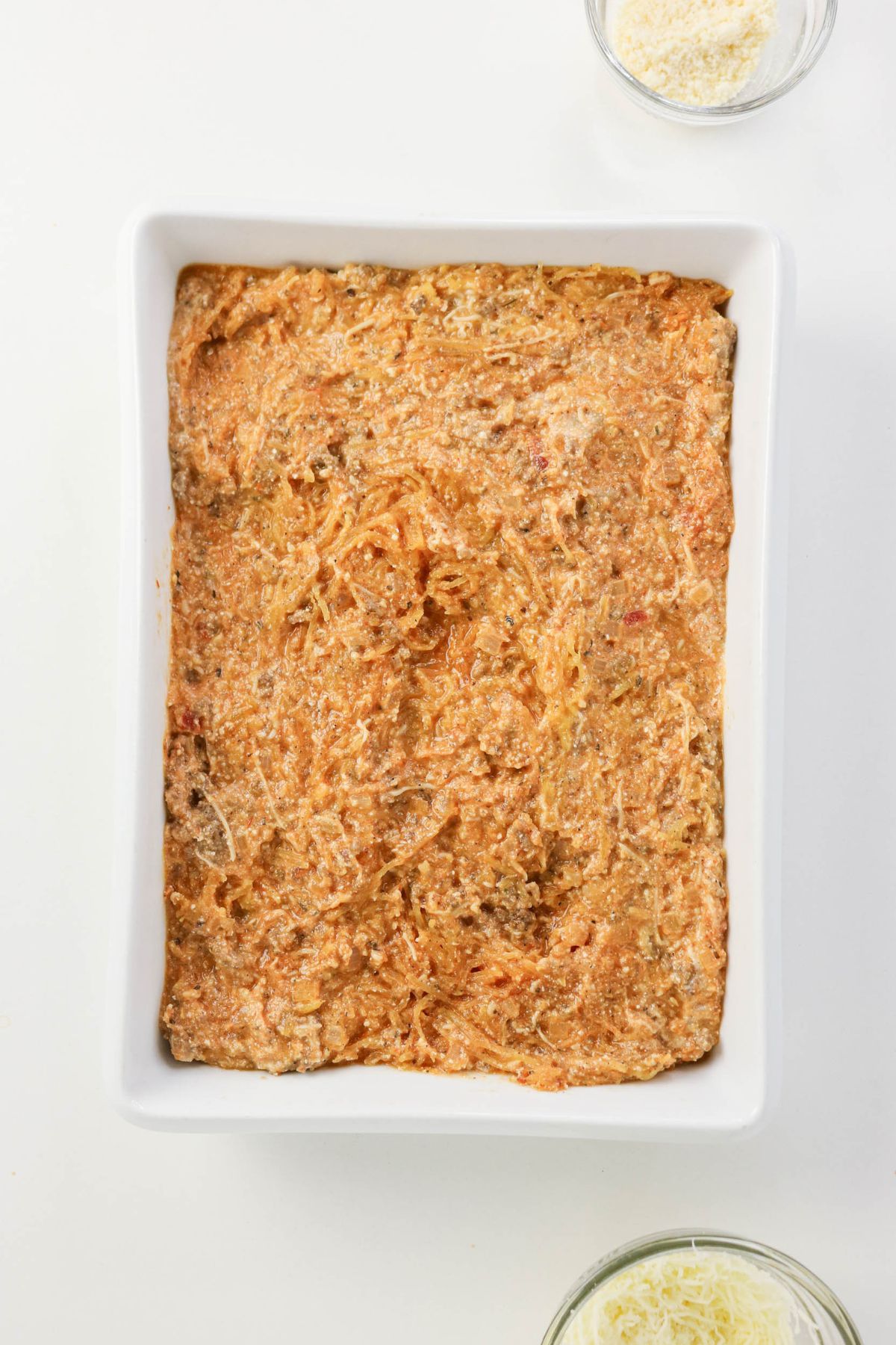 Rectangular white baking dish filled with an uncooked, mixed casserole mixture, with small bowls of shredded cheese nearby on a white surface.