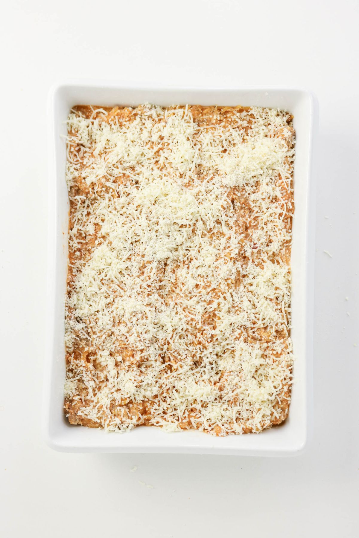 A rectangular white baking dish filled with a casserole topped with a generous layer of grated cheese, viewed from above on a white background.