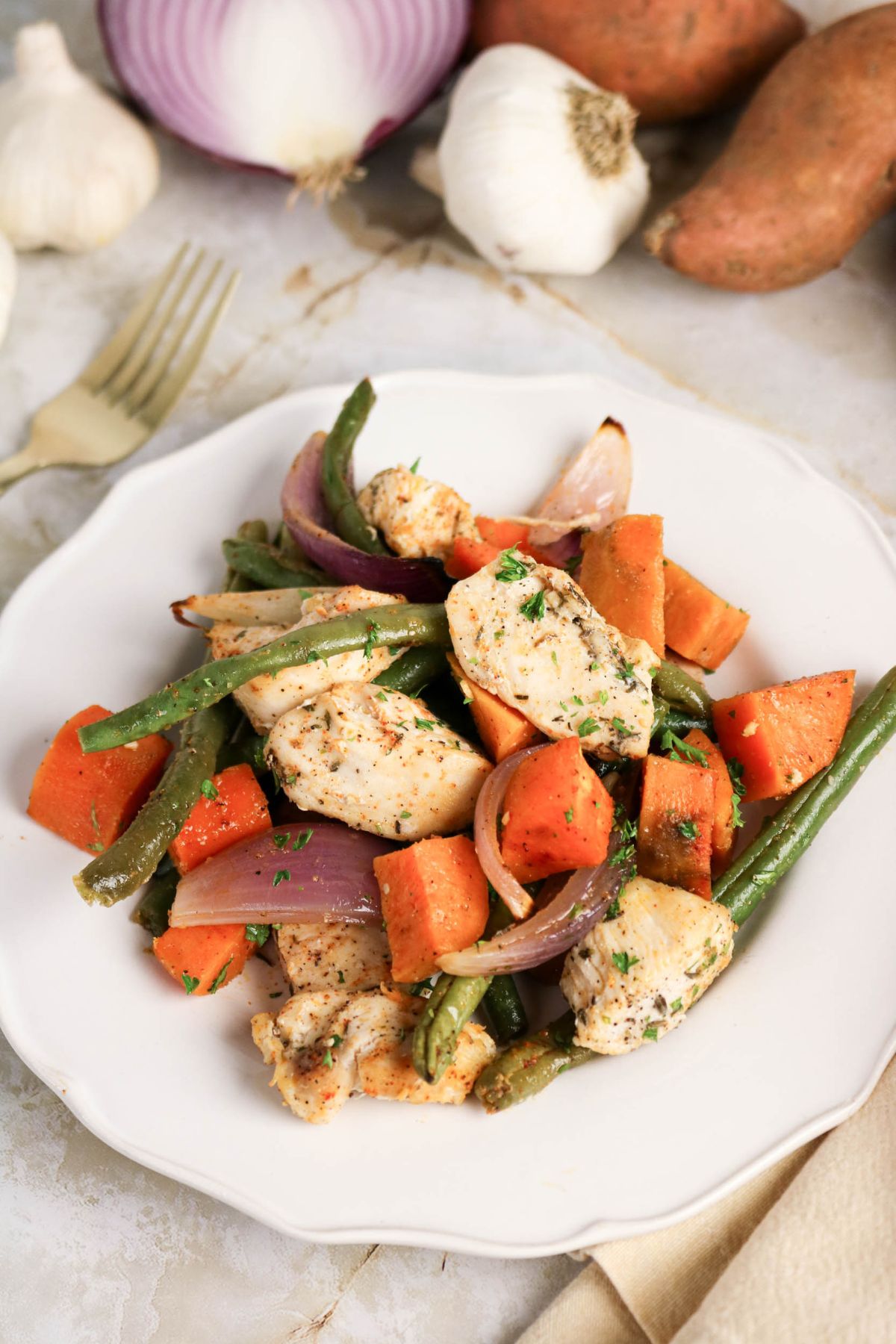 A white plate with cooked chicken pieces, green beans, sweet potatoes, and red onion chunks, garnished with herbs. Garlic, sweet potatoes, and a red onion are in the background.