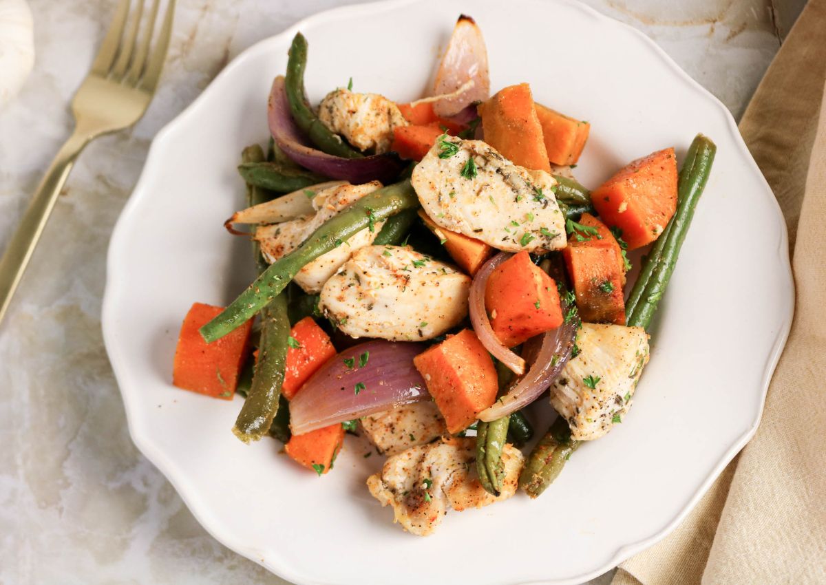A plate of cooked chicken breast, green beans, sweet potatoes, and red onions, garnished with herbs, sits on a white plate with a fork beside it.