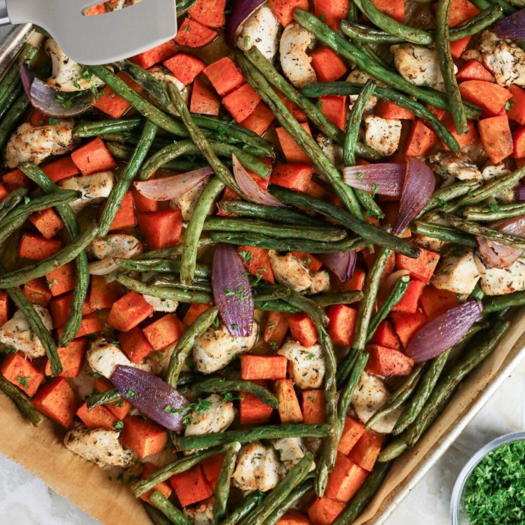Sheet pan with roasted green beans, sweet potatoes, red onions, and chicken pieces, all seasoned and baked, viewed from above.