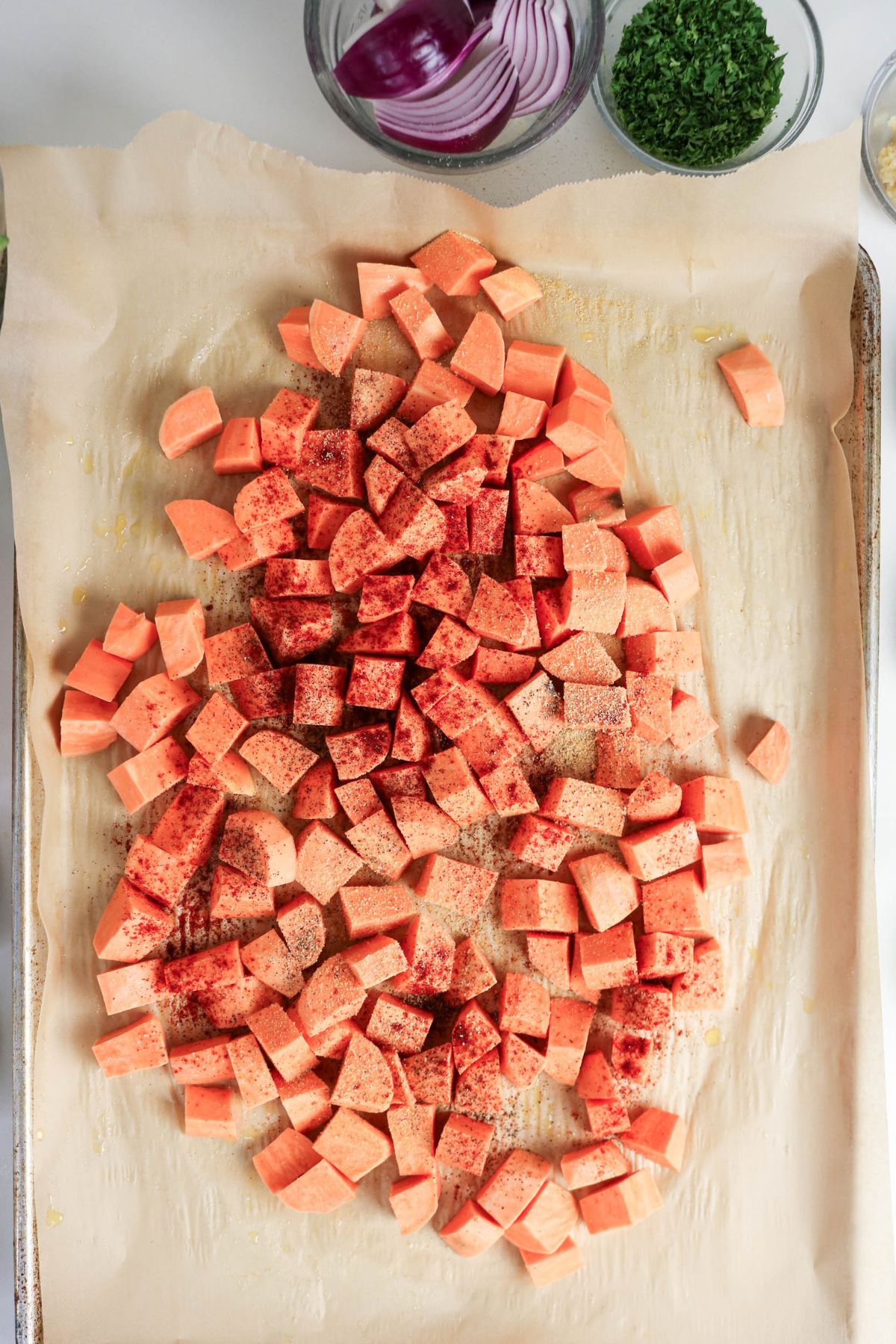 Diced sweet potatoes seasoned with spices are spread on a parchment-lined baking sheet, with bowls of red onion slices and chopped parsley visible in the background.
