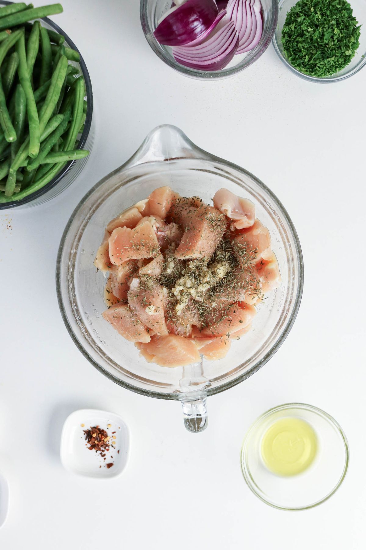 Raw cubed chicken breast in a glass bowl with herbs and spices, surrounded by green beans, red onion, parsley, oil, and red pepper flakes on a white surface.