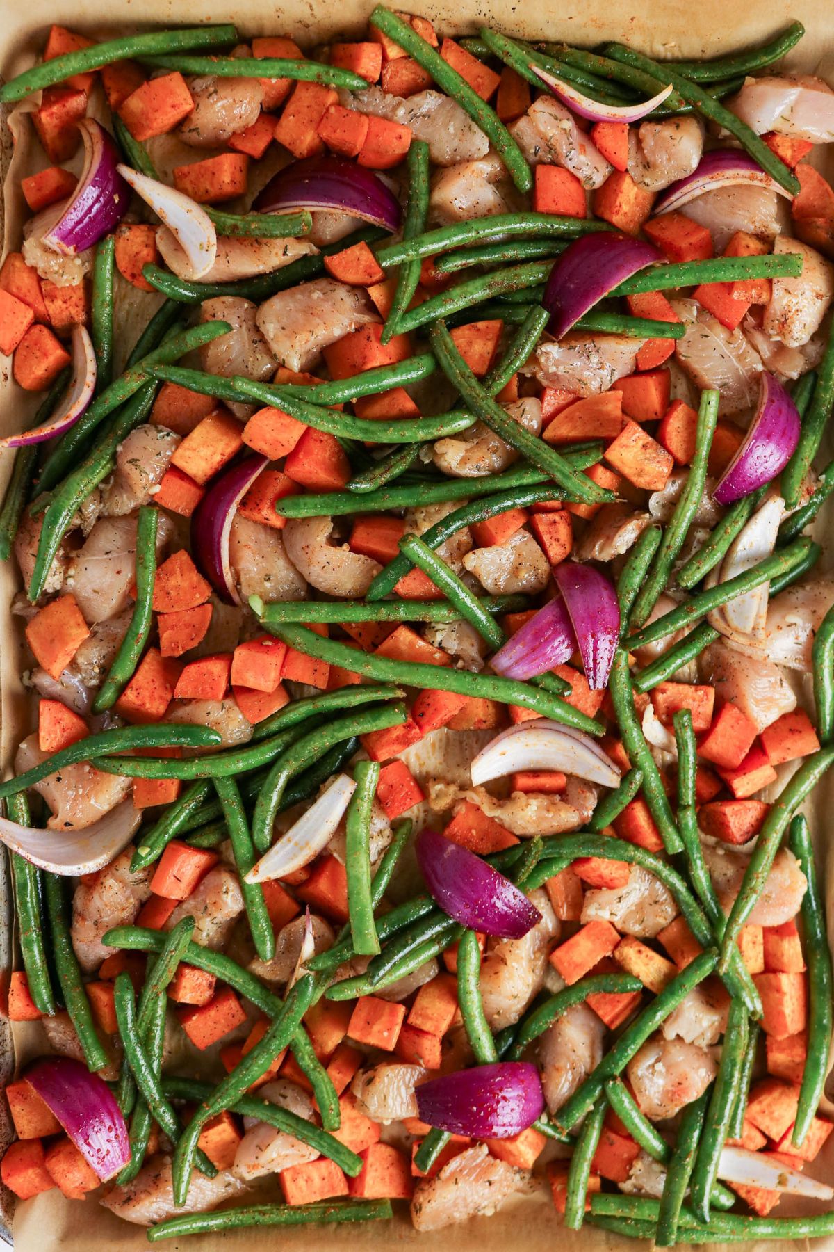 Sheet pan with raw chicken pieces, diced sweet potatoes, green beans, and red onion wedges, all seasoned and ready for roasting.