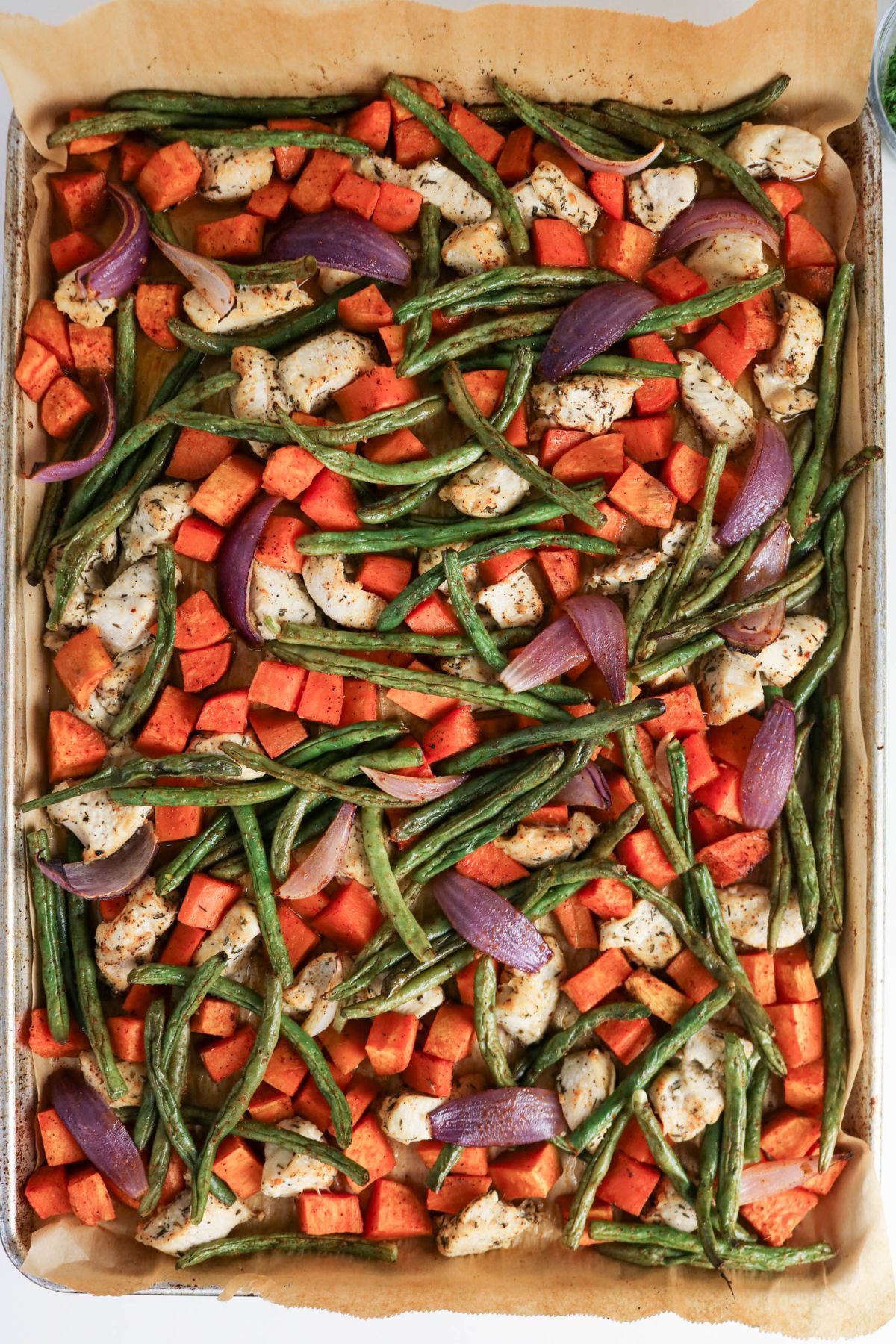 Sheet pan with roasted green beans, diced sweet potatoes, red onion wedges, and pieces of seasoned chicken on parchment paper.