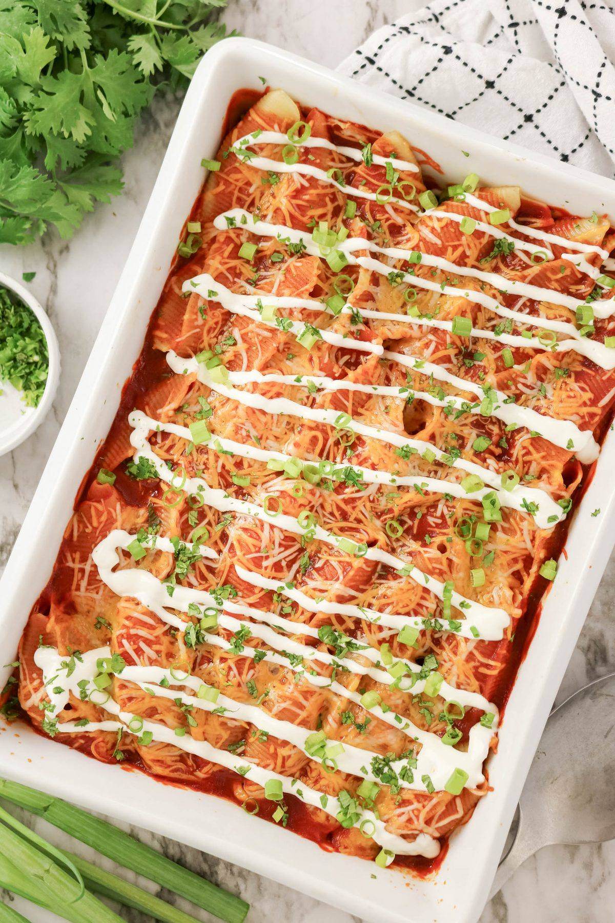 A rectangular baking dish filled with enchiladas topped with melted cheese, sour cream drizzle, and chopped green onions. Fresh cilantro and a kitchen towel are nearby.