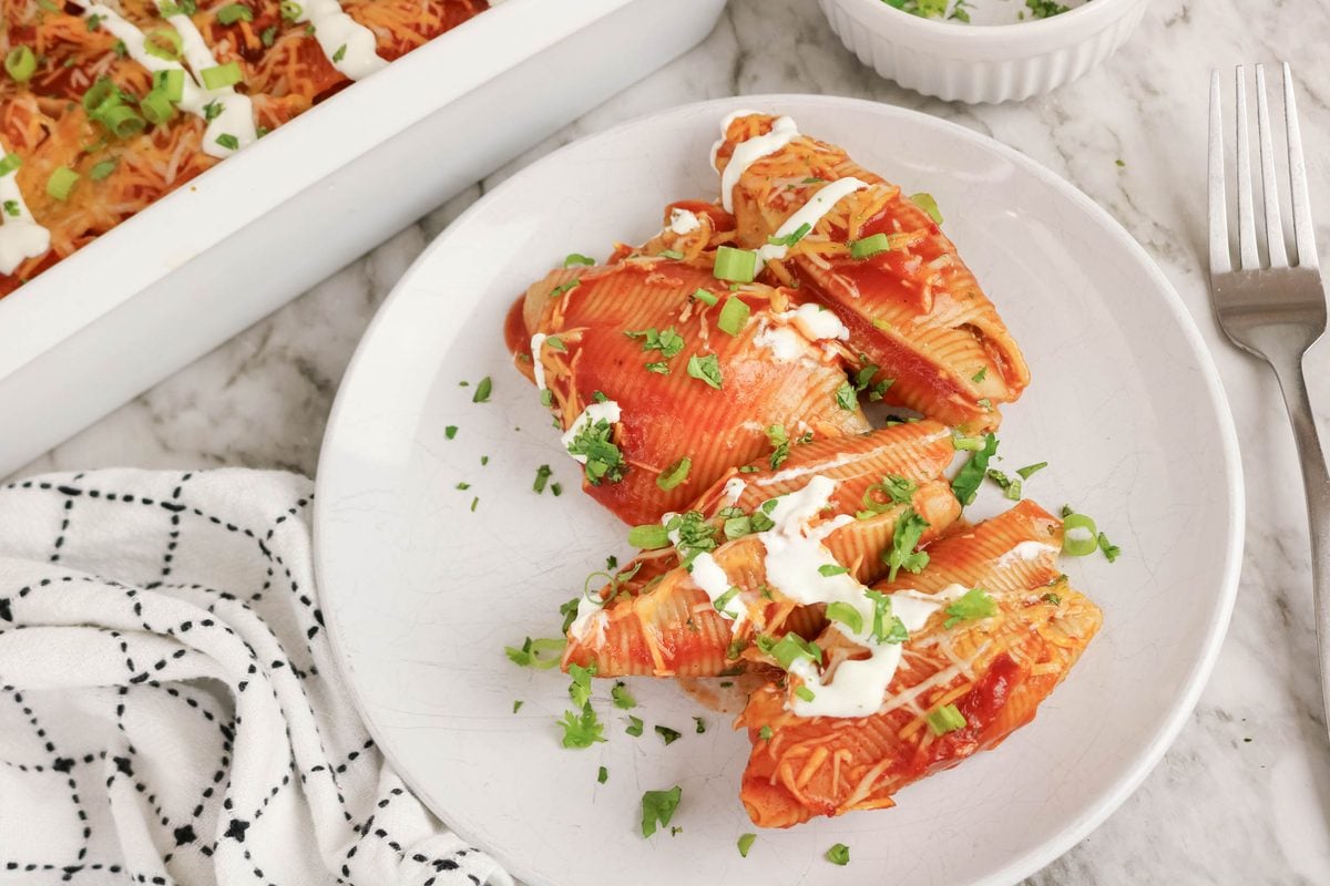 Three stuffed pasta shells topped with tomato sauce, shredded cheese, sour cream, and chopped green onions are served on a white plate next to a fork and a black-and-white napkin.
