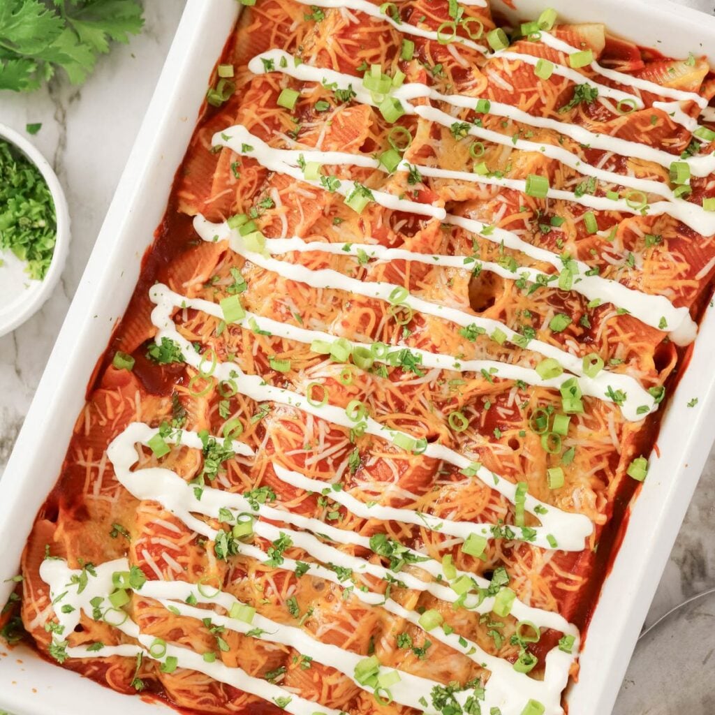 A baking dish of enchiladas topped with shredded cheese, chopped green onions, cilantro, and drizzled sour cream, with extra herbs in a bowl on the side.