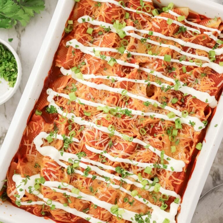 A baking dish of enchiladas topped with shredded cheese, chopped green onions, cilantro, and drizzled sour cream, with extra herbs in a bowl on the side.