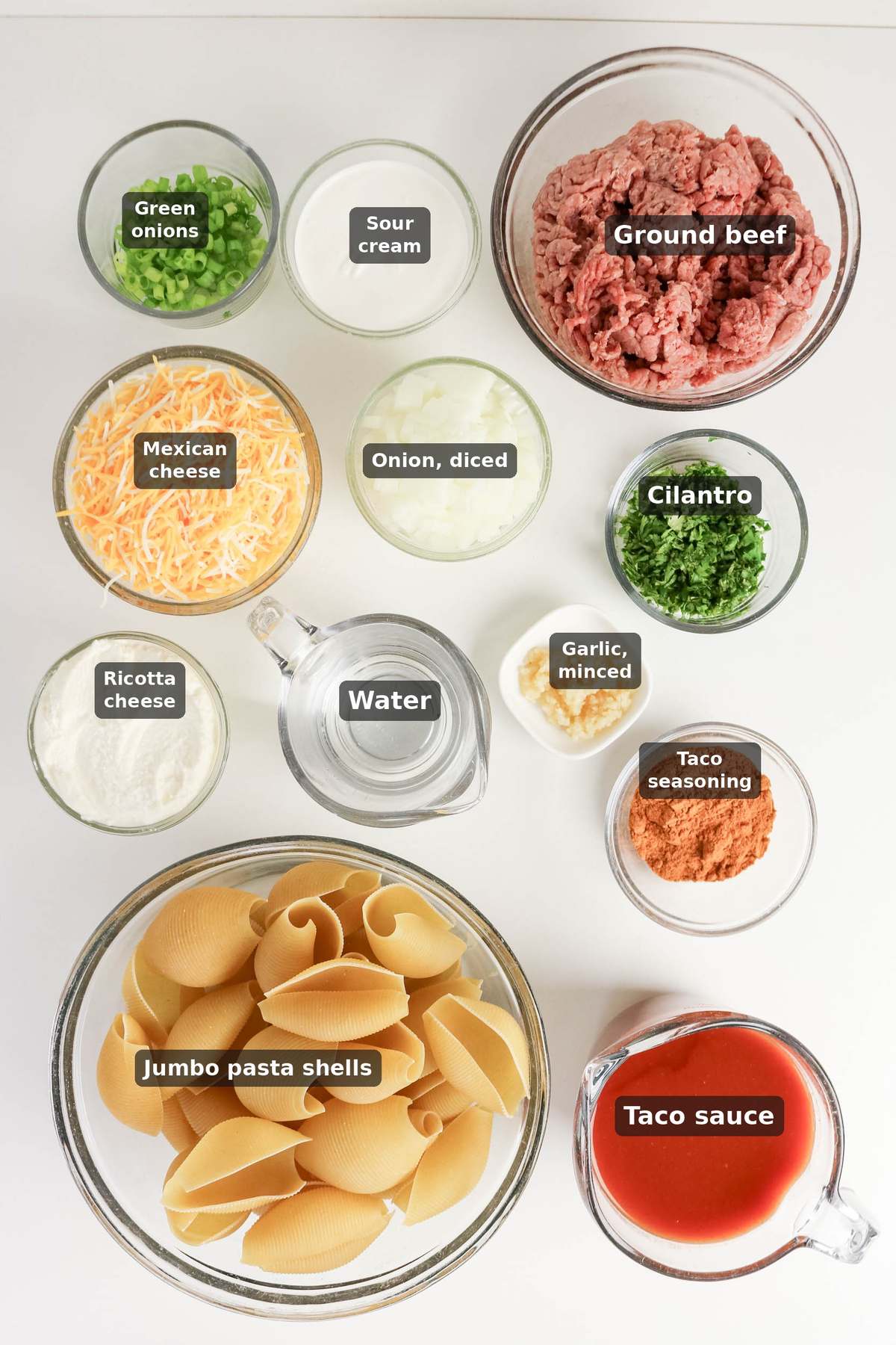 Top-down view of labeled ingredients in bowls for taco stuffed shells: ground beef, pasta shells, cheese, cilantro, onions, garlic, sour cream, ricotta, taco seasoning, water, and taco sauce.