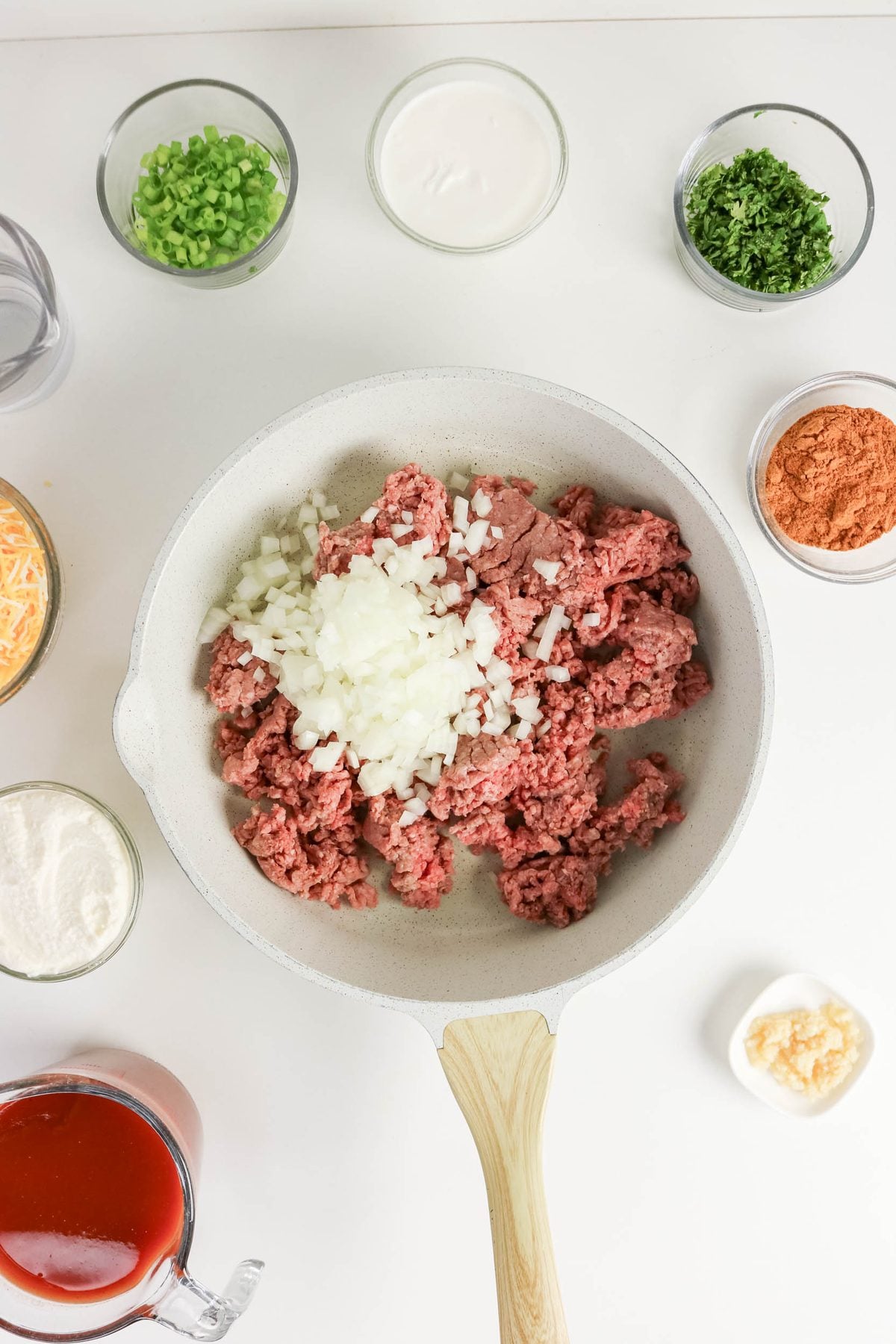 A skillet with raw ground beef and chopped onions, surrounded by bowls of green onions, parsley, shredded cheese, sauce, cream, spices, tomato sauce, flour, and minced garlic.