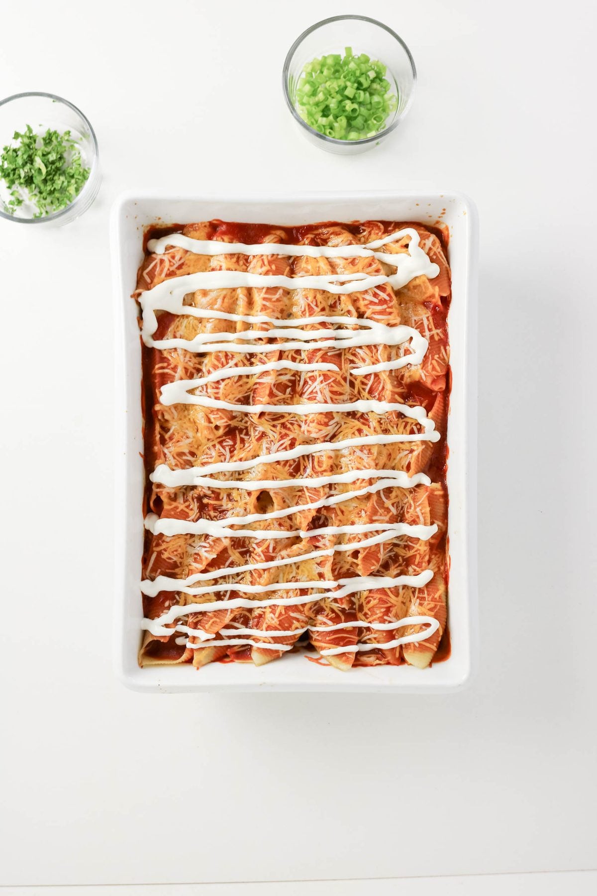 A rectangular dish of enchiladas topped with melted cheese and drizzled with sour cream, with two bowls of chopped green onions on the side.