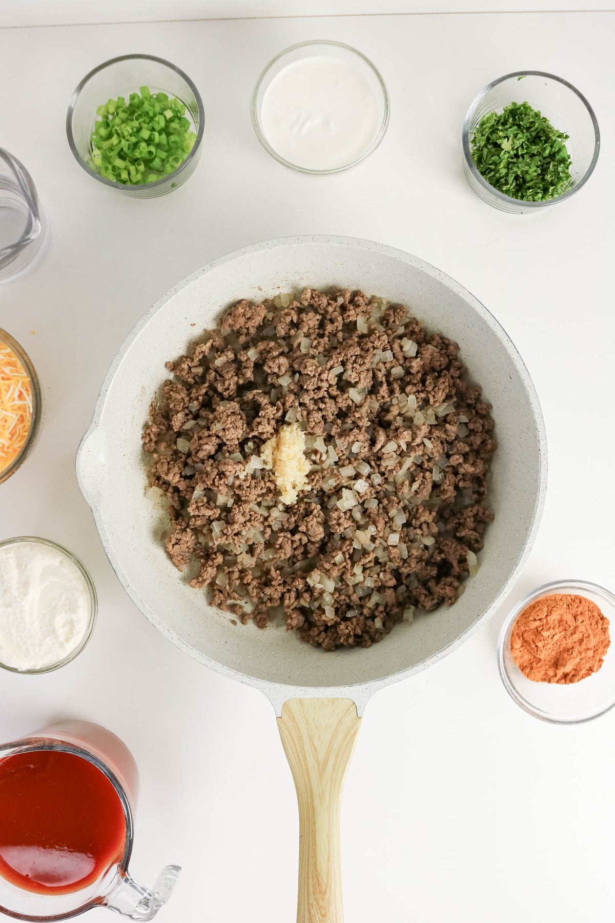 White skillet with cooked ground beef and onions in the center, surrounded by bowls of chopped green onions, grated cheese, parsley, sour cream, spices, and a measuring cup of red sauce.