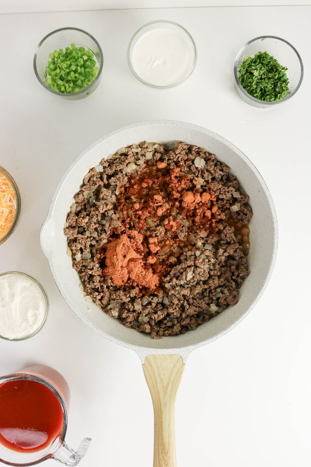A skillet with cooked ground beef and spices is surrounded by bowls of shredded cheese, sour cream, green onions, herbs, tomato sauce, and other ingredients on a white surface.