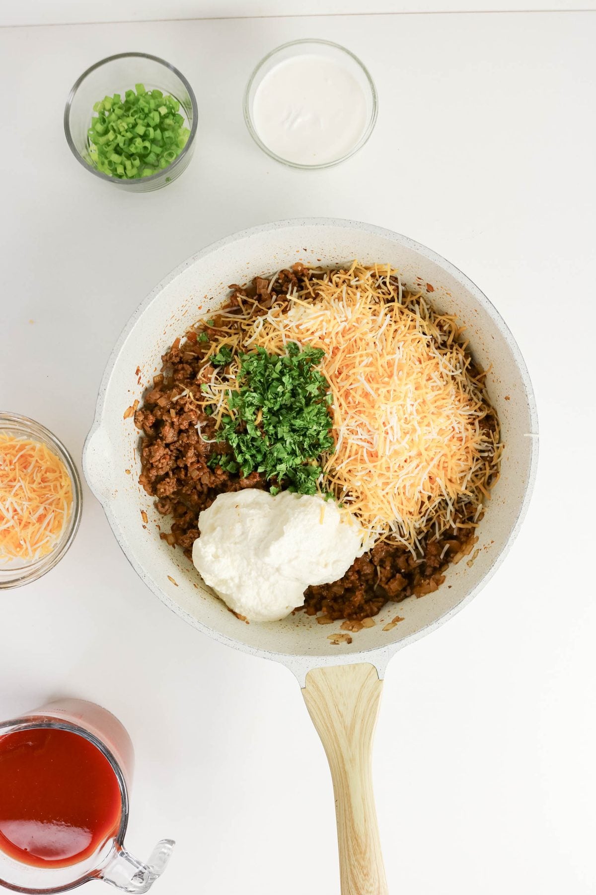 A white skillet with cooked ground beef, shredded cheese, sour cream, and chopped herbs. Surrounding the skillet are small bowls of green onions, shredded cheese, sour cream, and a cup of red sauce.