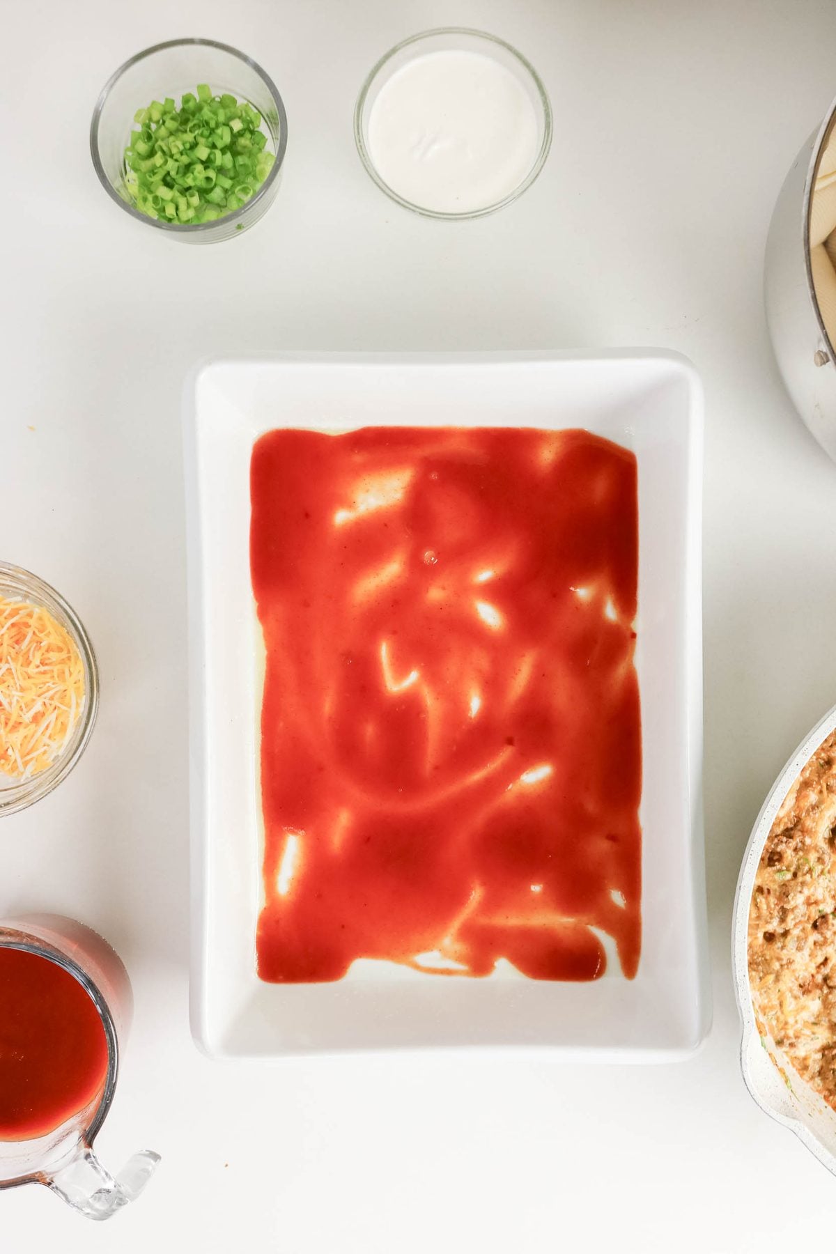 A rectangular white dish with red sauce spread on the bottom, surrounded by bowls of cheese, green onions, sour cream, more sauce, and a pot with a mixture.
