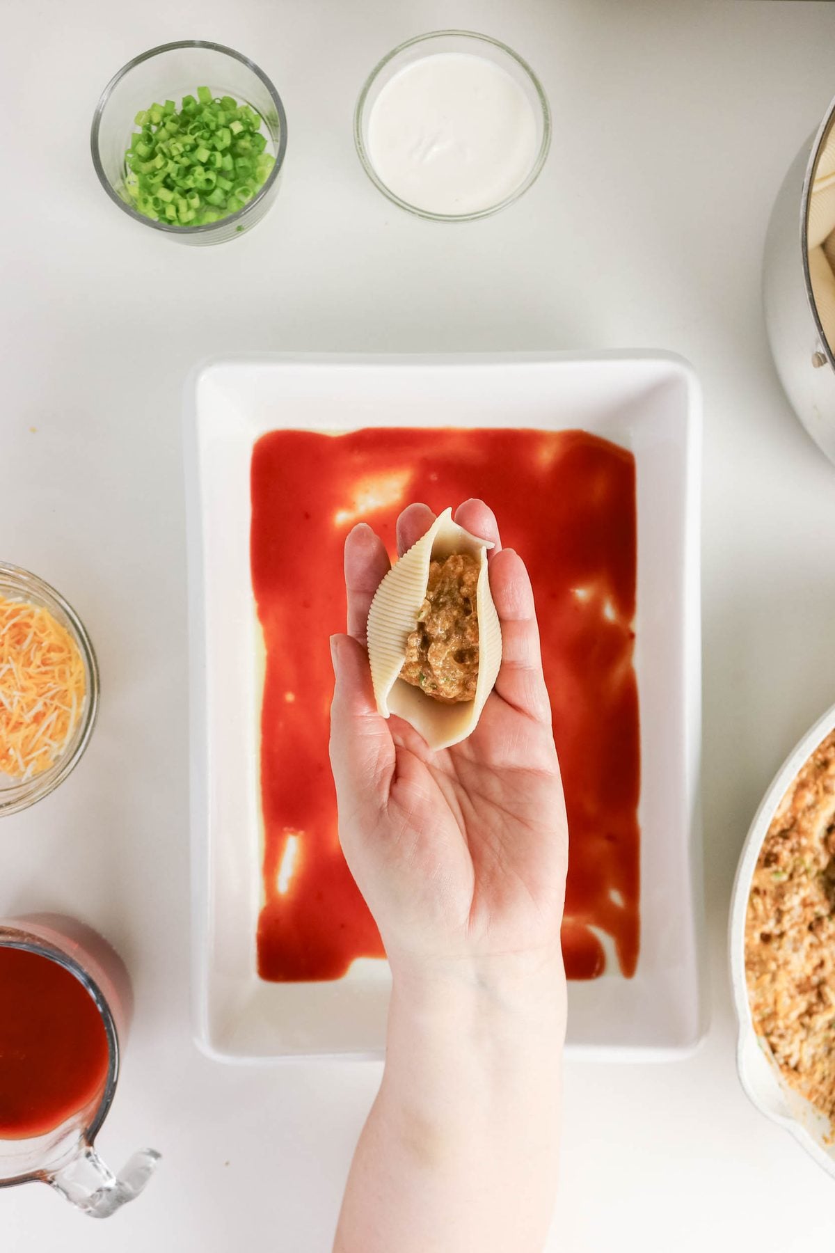 A hand holds a pasta shell filled with meat mixture above a baking dish lined with tomato sauce, surrounded by bowls of ingredients.
