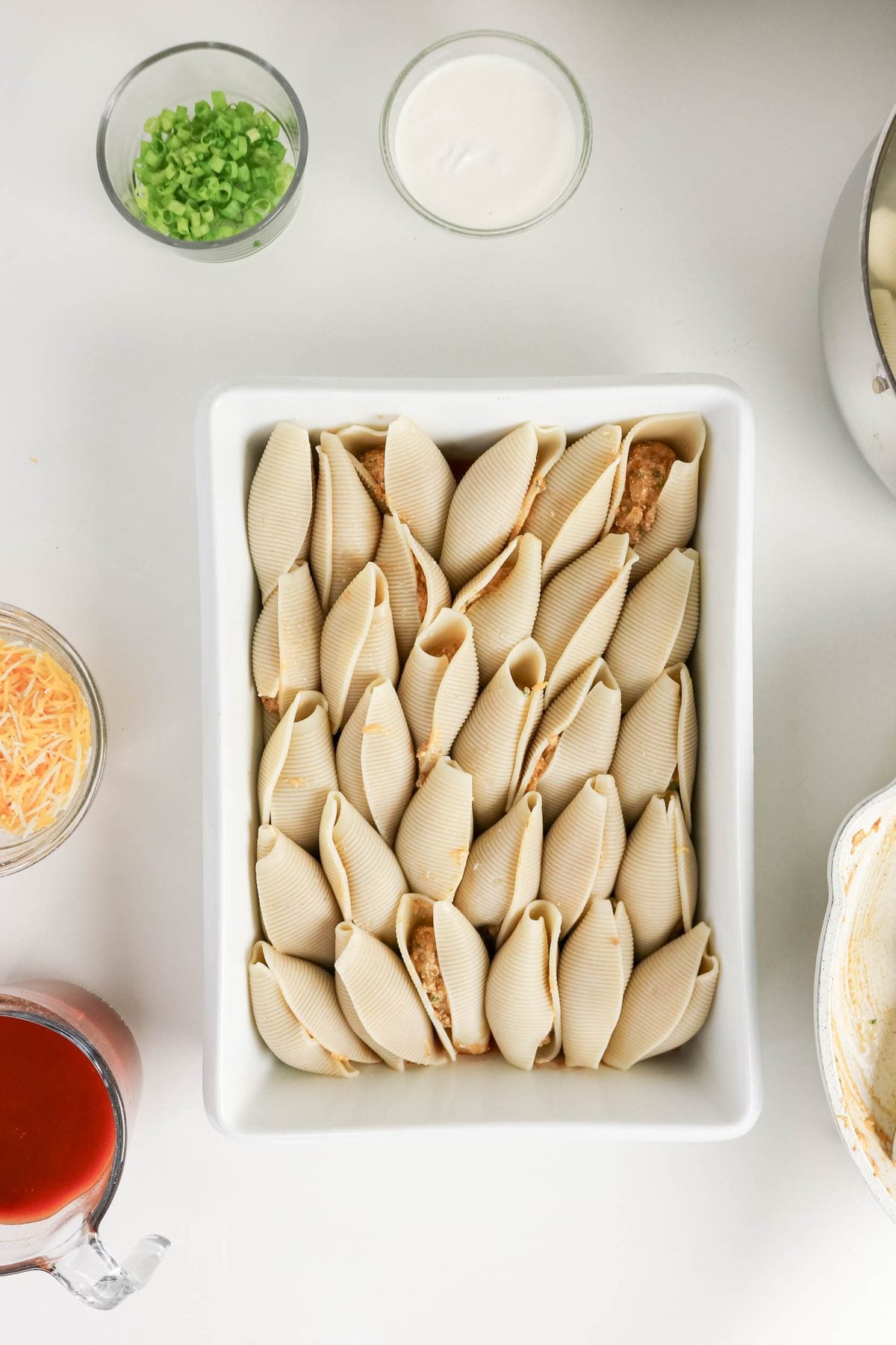 Uncooked stuffed pasta shells arranged in a white baking dish, surrounded by bowls of shredded cheese, chopped green onions, sauce, and a measuring cup of tomato sauce on a white surface.