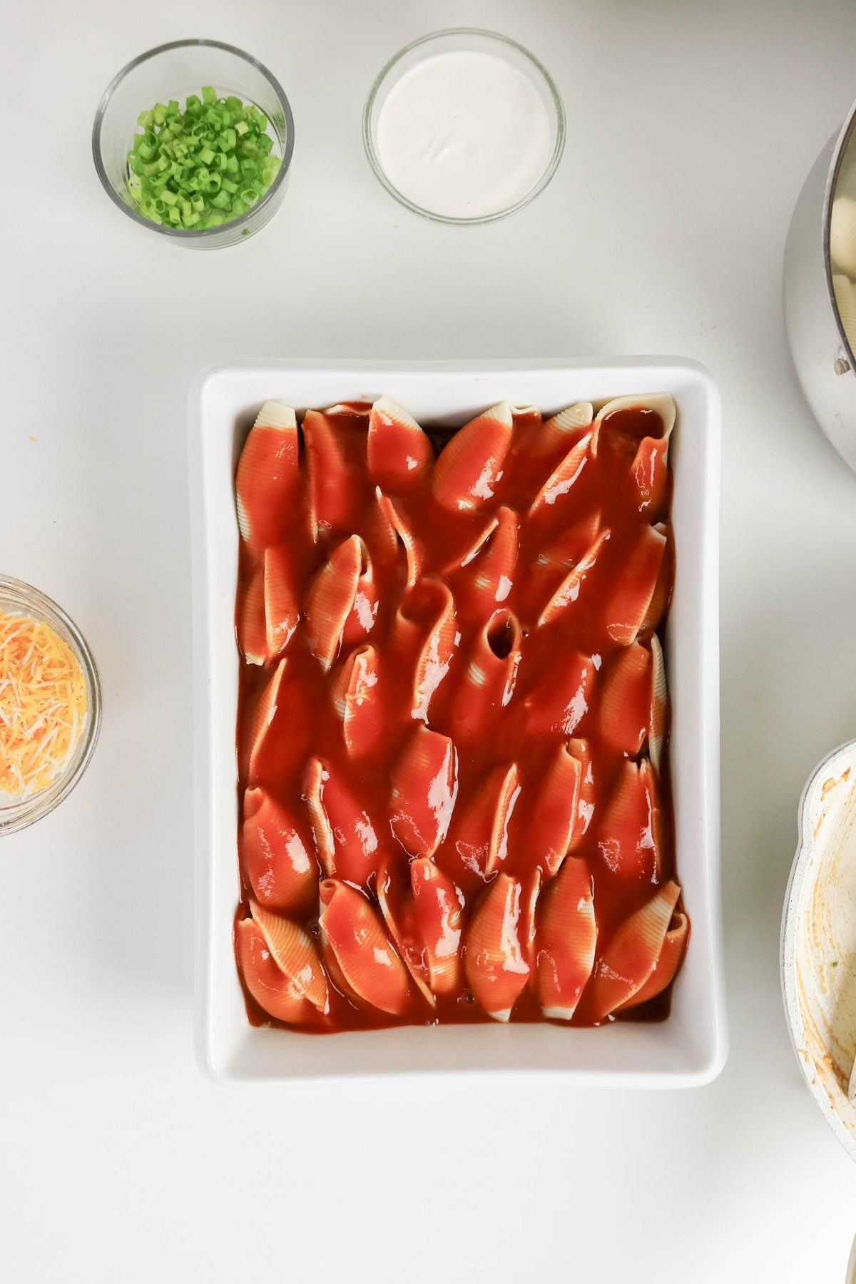 A white baking dish filled with large pasta shells covered in red sauce, surrounded by bowls of green onions, shredded cheese, and sour cream on a white surface.