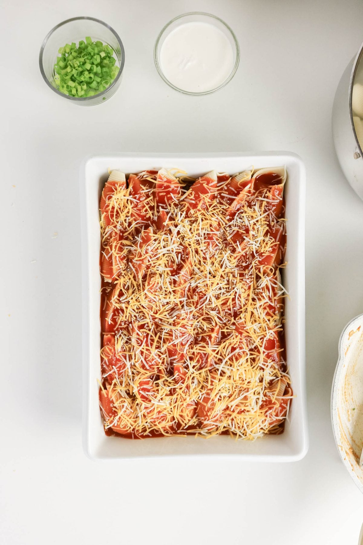 Rectangular dish filled with uncooked enchiladas topped with shredded cheese, next to bowls of chopped green onions and sour cream on a white surface.