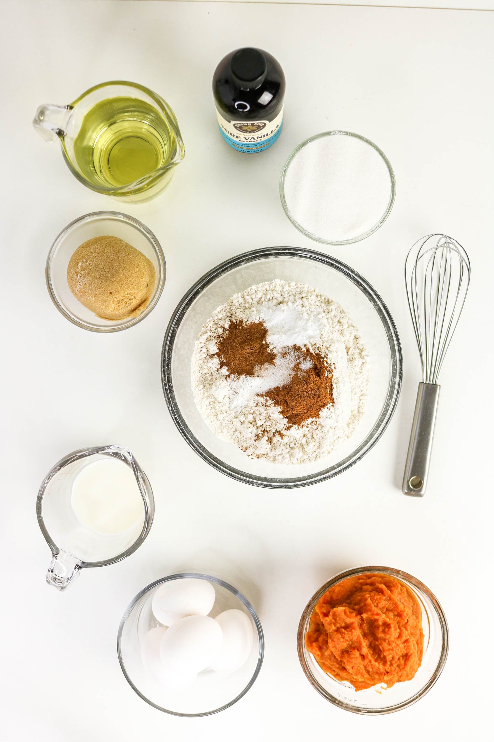 Ingredients for a pumpkin bundt cake, including a bowl of flour with spices, eggs, pumpkin puree, milk, oil, vanilla, and sugar, arranged around a whisk on a white surface.