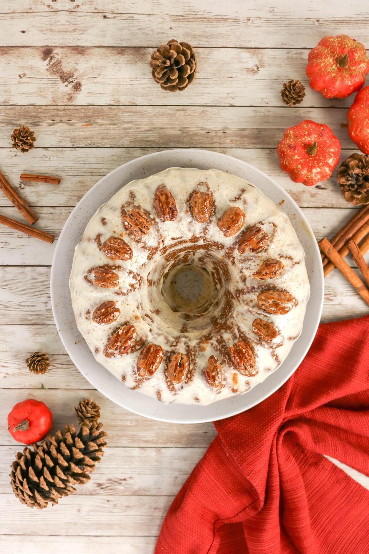A bundt cake topped with white frosting and pecans on a white plate, surrounded by pinecones, cinnamon sticks, and small decorative pumpkins on a wooden surface.