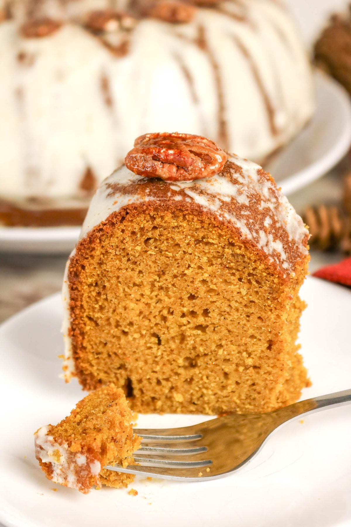 A slice of glazed pumpkin bundt cake topped with a pecan, with a fork holding a small piece of the cake on a white plate.