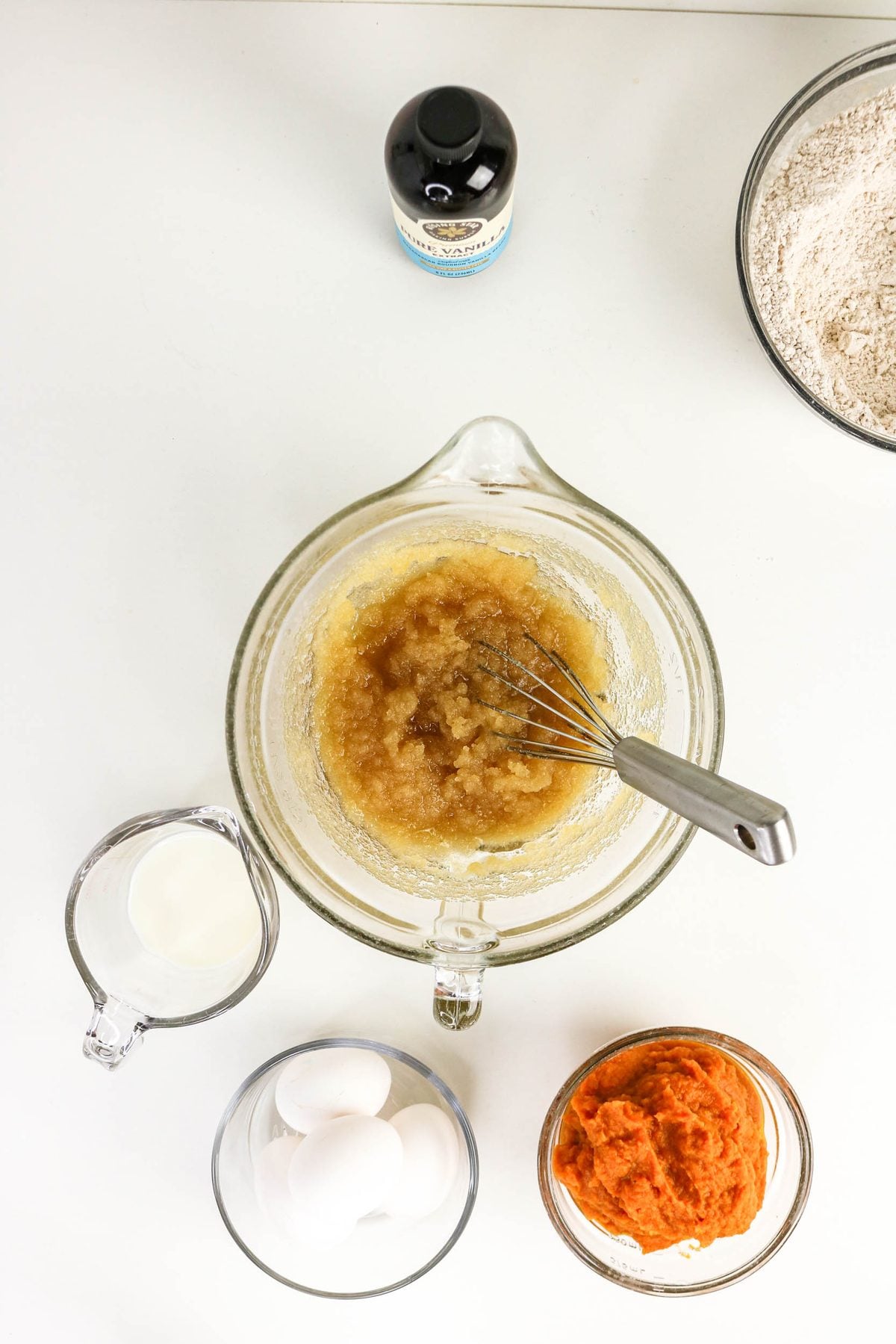 A glass mixing bowl with whisked sugar and butter, surrounded by a bottle of vanilla, flour, milk, eggs, and pumpkin puree on a white surface.
