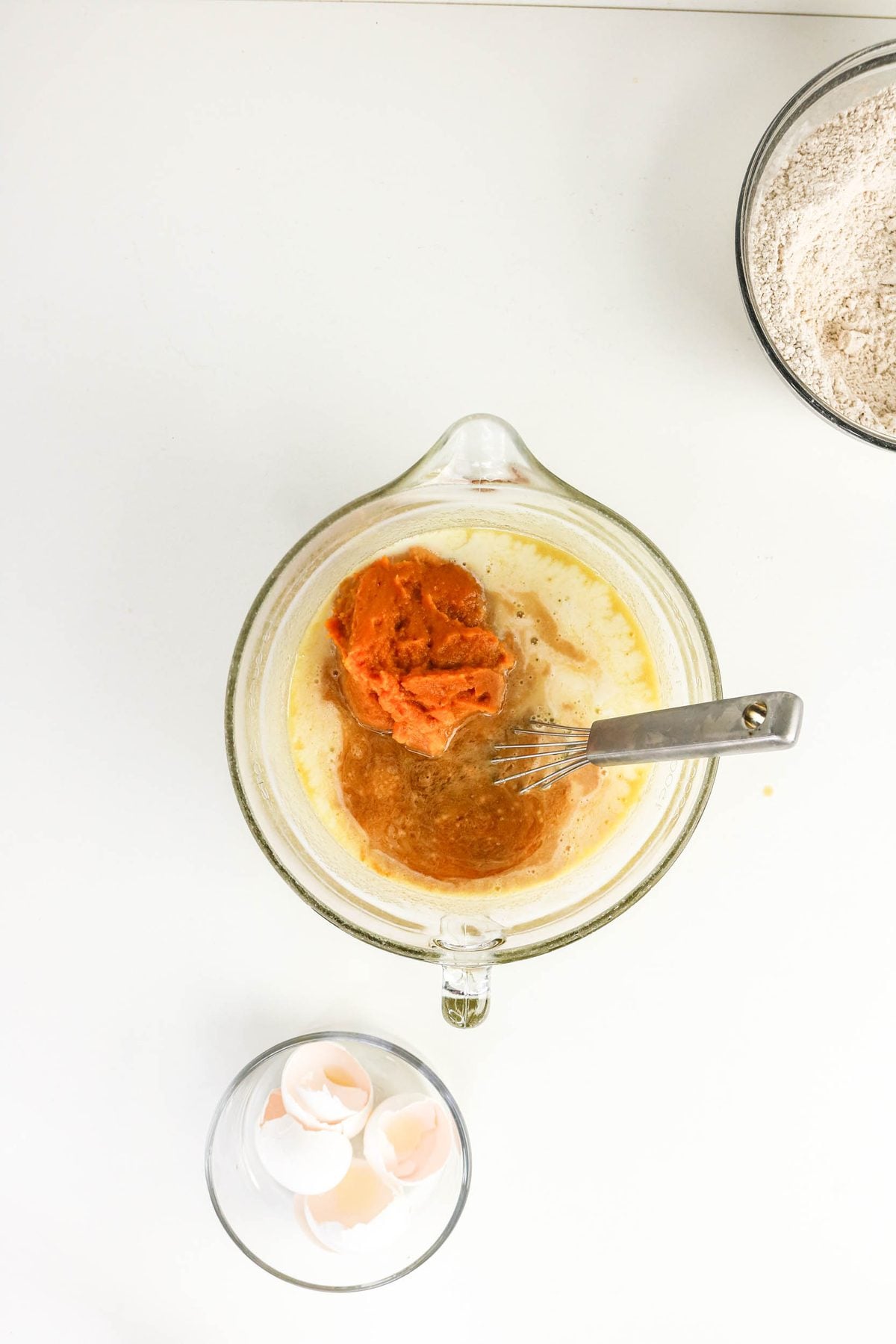 Pumpkin puree, eggs, and liquid ingredients in a glass measuring cup with a whisk, next to a bowl of flour and a bowl of eggshells.