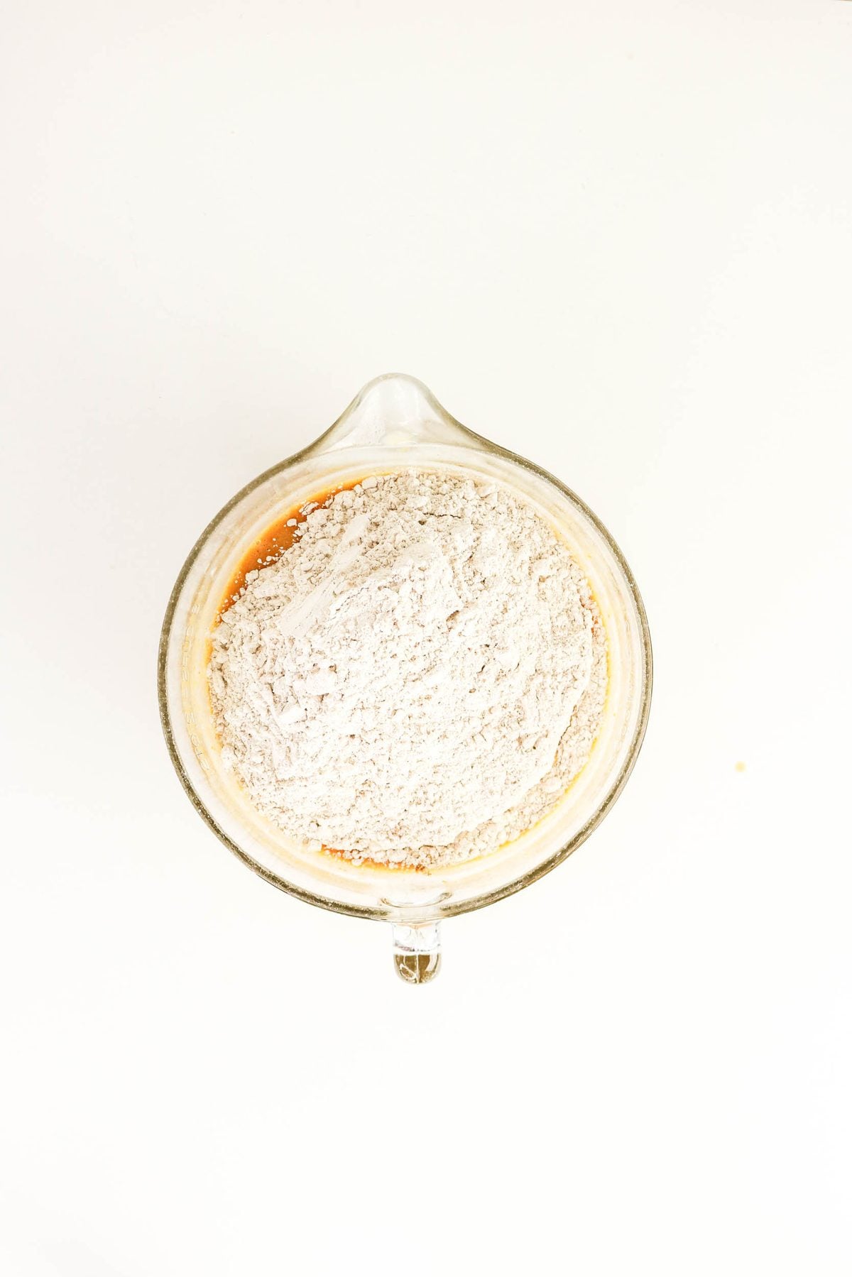 A pile of flour resting on top of liquid ingredients in a glass measuring cup.
