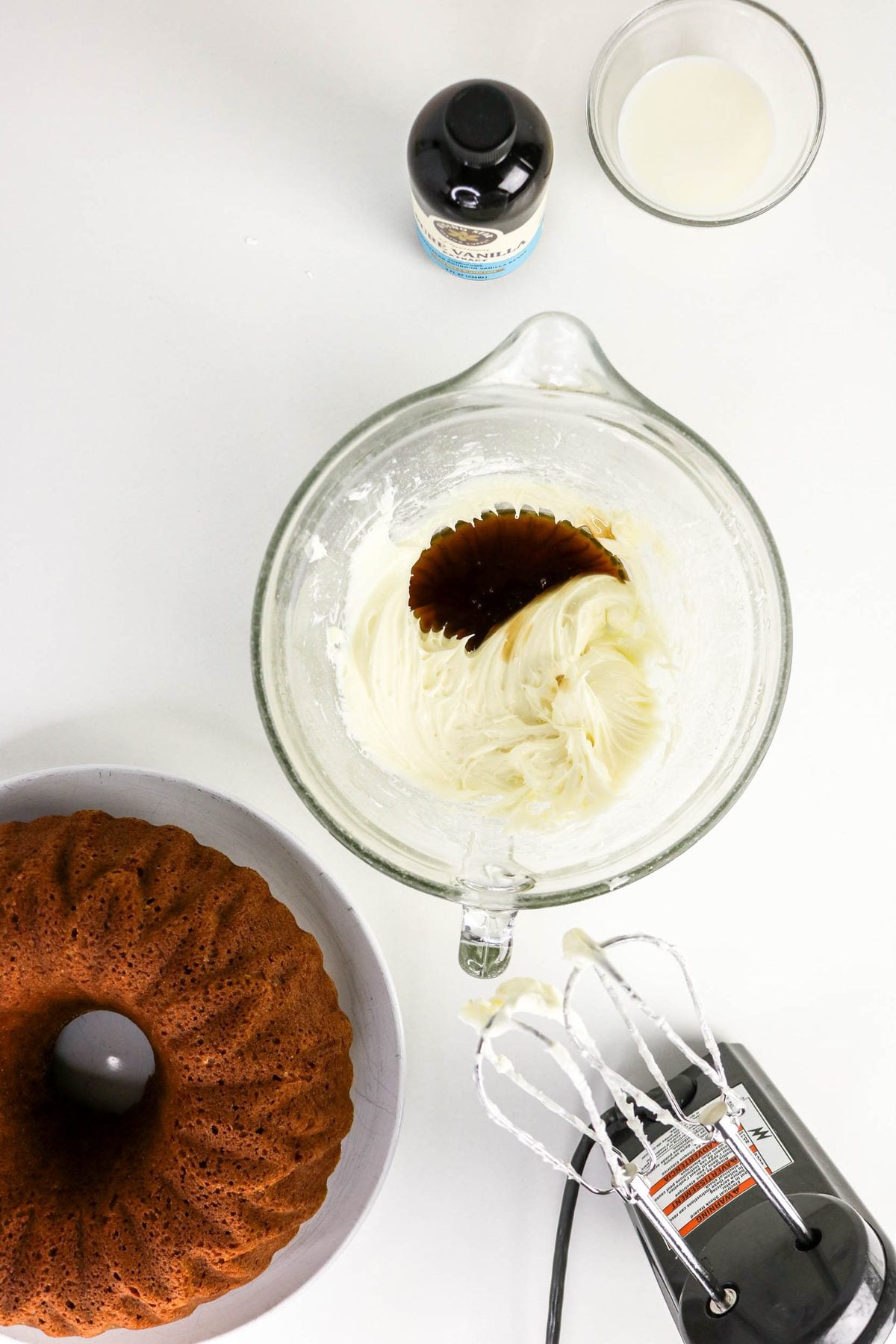 A glass bowl containing white frosting being mixed with a splash of dark vanilla extract, next to a bundt cake, a bottle of vanilla, a small bowl of milk, and an electric hand mixer.