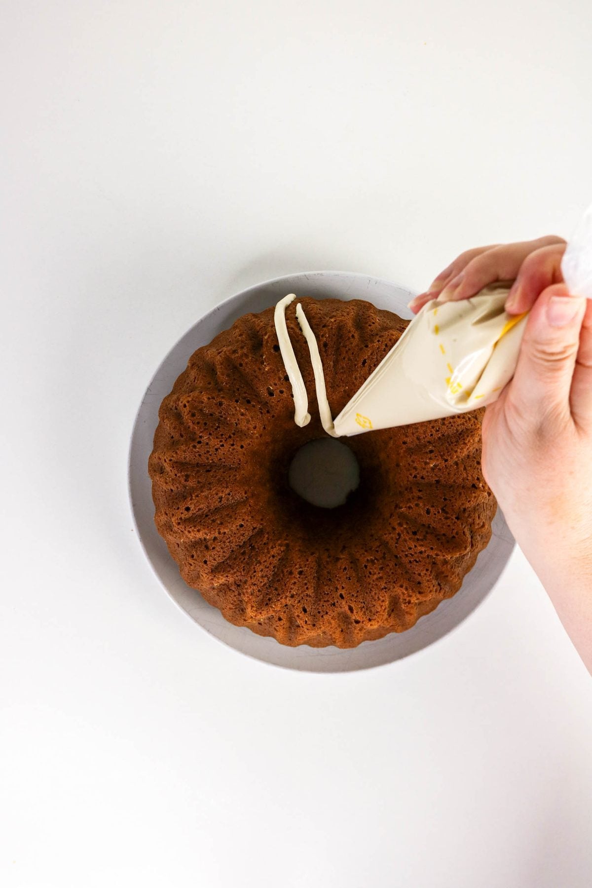 A hand using a piping bag to drizzle icing onto a ring-shaped bundt cake on a plate.