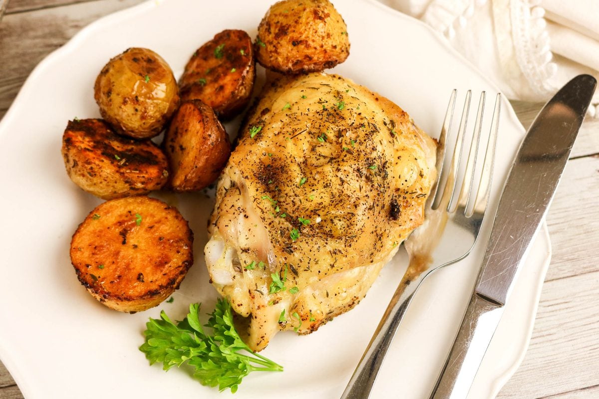 A roasted chicken thigh with herbs served with halved roasted potatoes and a sprig of parsley on a plate with a fork and knife.